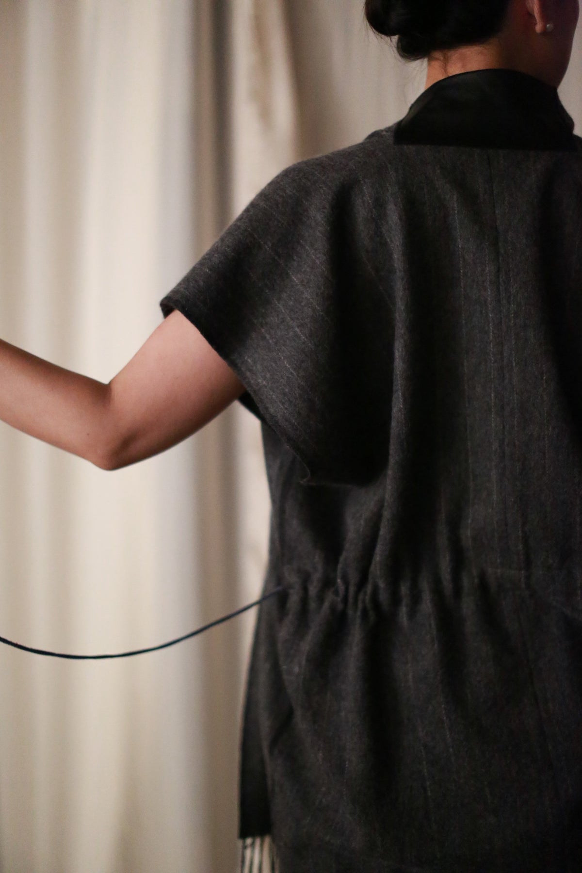 A person with short dark hair, seen from behind, wears the Men's Scarf Vest in Charcoal—handmade in San Francisco. The loose-fitting, short-sleeve vest is paired with a thin black cord held in their right hand, against light curtains.