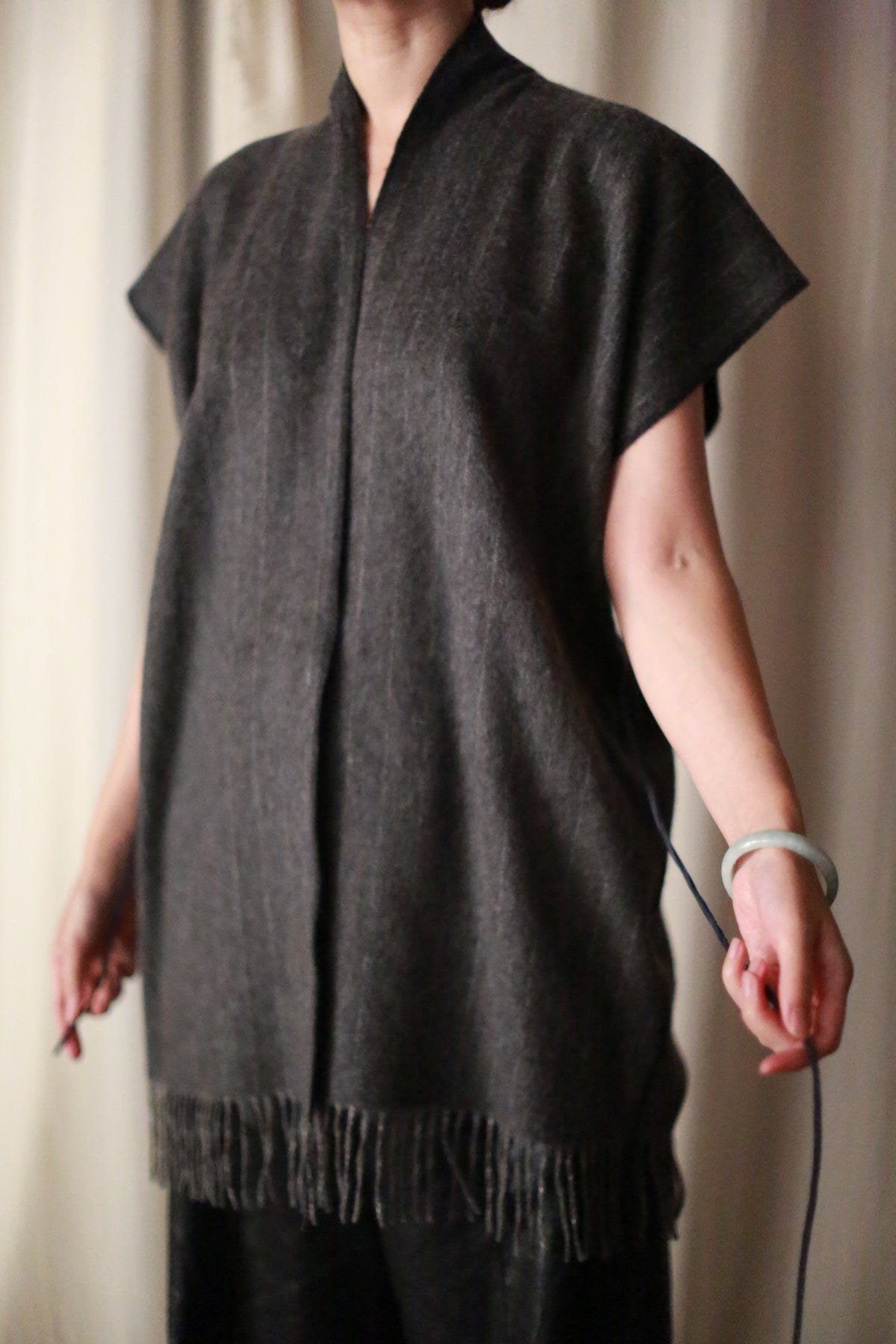 A person indoors wears the Men's Scarf Vest in Charcoal—a loose, dark gray vest with fringe and short sleeves—styled with a bracelet and featuring zero waste design. Their face is not visible.
