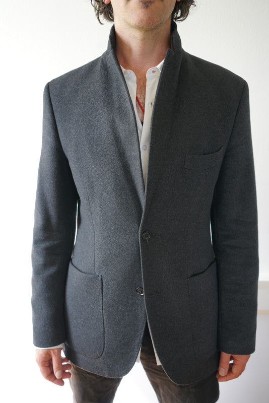 A person modeling the Men's Cashmere Blazer in charcoal gray, designed with a tailored European fit, is crafted from soft cashmere and layered over a white dress shirt. The blazer features a high collar and two-button fastening, and they stand against a white backdrop, shown from the neck down without their face visible.