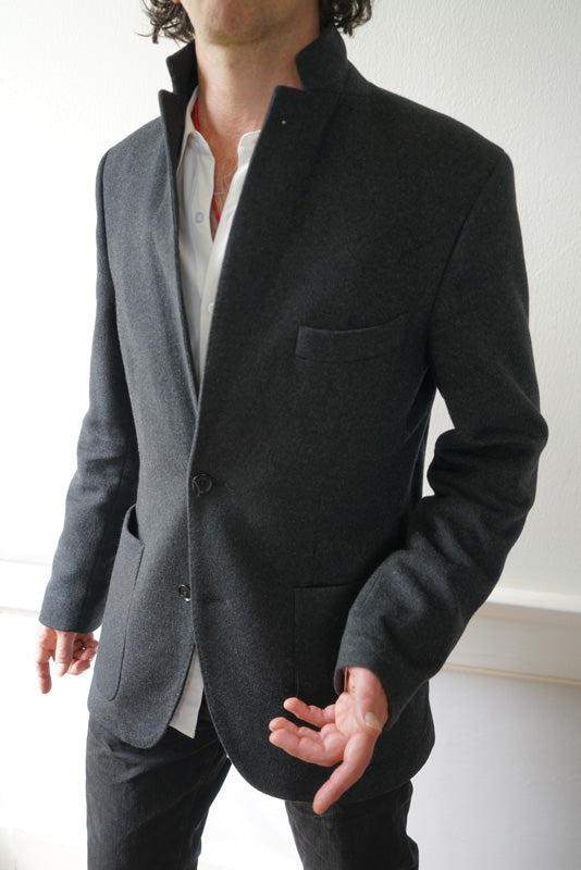 A person is standing indoors, sporting the Men's Cashmere Blazer in charcoal gray with a European fit over a white shirt. This meticulously handcrafted blazer highlights luxurious cashmere, complete with a chest pocket and two front buttons. One hand is casually tucked into their pocket while the other is slightly lifted, though their head is cropped out of the frame.