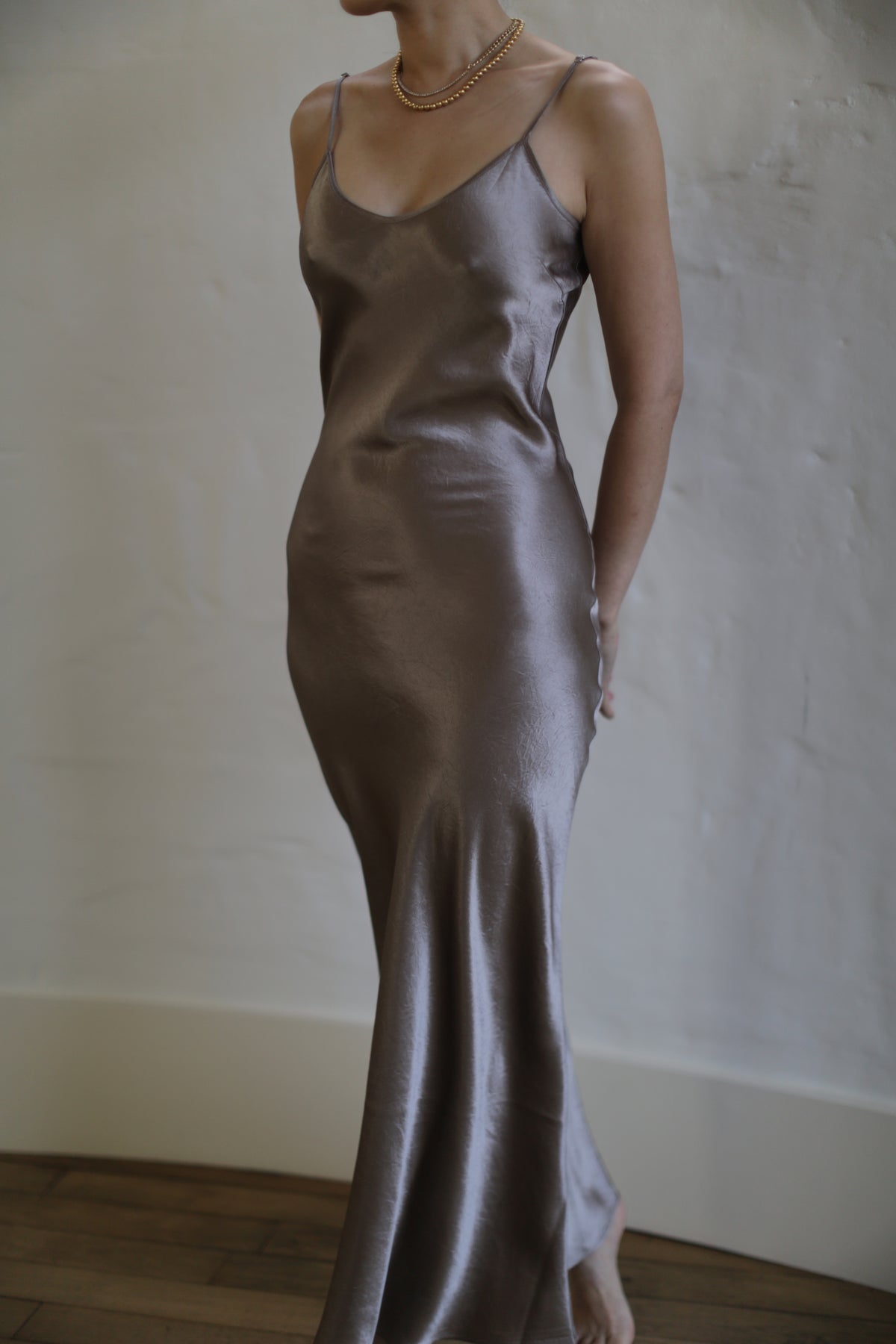A person is wearing a Liquid Slip Dress Long | Mink made from plant-derived material, featuring a satin, silvery-gray slip design with thin straps. They are also wearing a thin gold necklace. The background is plain with light-colored walls, and the floor is wooden. The image is taken indoors with soft lighting.