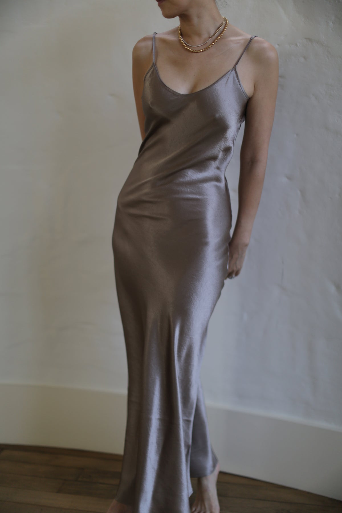 A person wearing a Liquid Slip Dress Long | Mink with thin straps is standing against a textured, light-colored wall. The bias cut of the dress beautifully accentuates their figure, complemented by a layered gold necklace. The lighting highlights the shine and smooth texture of the ensemble.