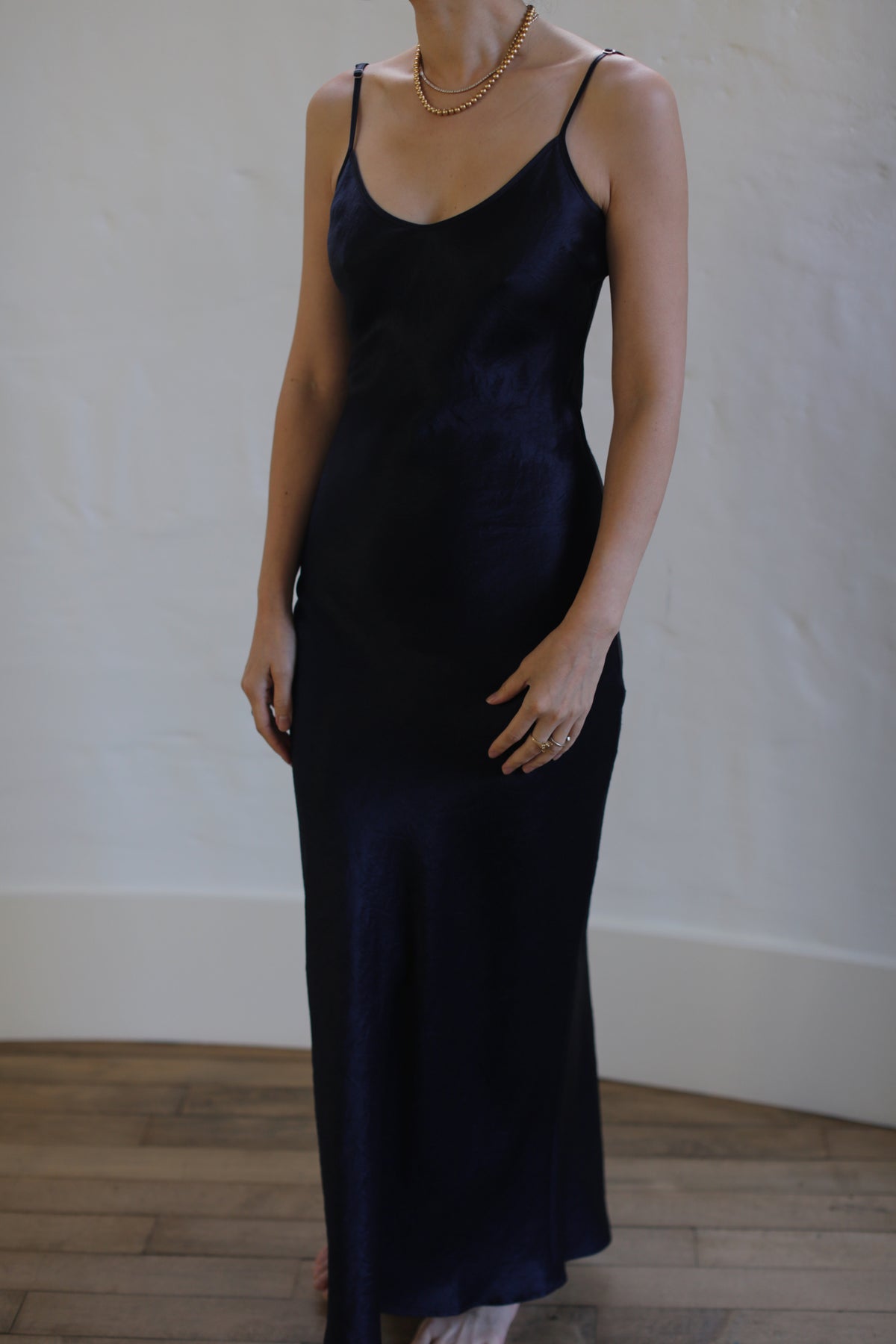 A person wearing a sleek, sleeveless Liquid Slip Dress Long | Midnight stands indoors against a neutral wall. They are accessorized with a layered gold necklace and a ring on their left hand. The wooden floor adds a warm contrast to the minimal background.