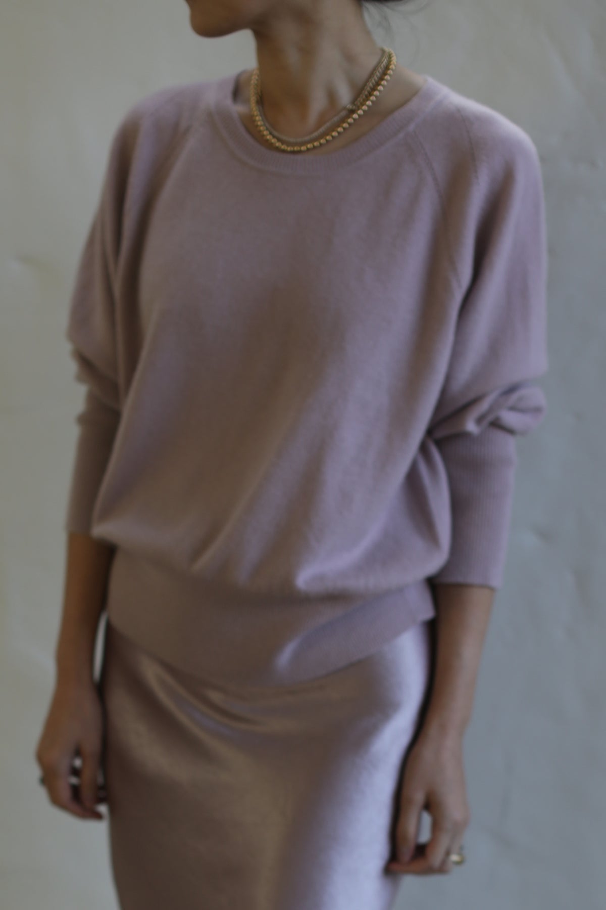A person is dressed in a light purple, loose-fitting, long-sleeved Raglan Crewneck Cashmere Sweater | Peony and matching silky skirt. They are also wearing a gold necklace. The photo is taken from the chest down, and the background is plain and neutral.
