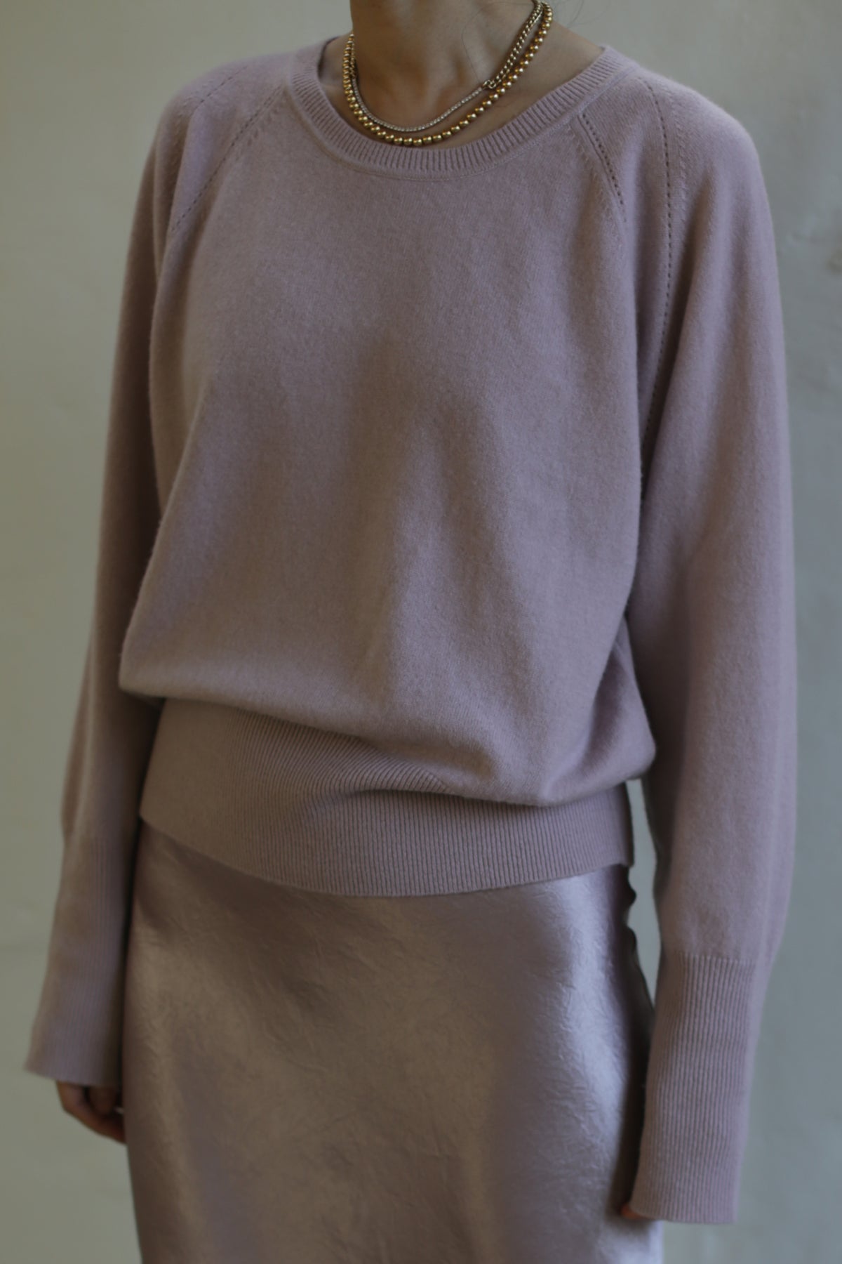A person wearing a light purple Raglan Crewneck Cashmere Sweater | Peony and a metallic skirt. The upper part of their body is shown, with the head cropped out of the image. They are also wearing a gold necklace, adding an elegant touch to this chic wardrobe staple.