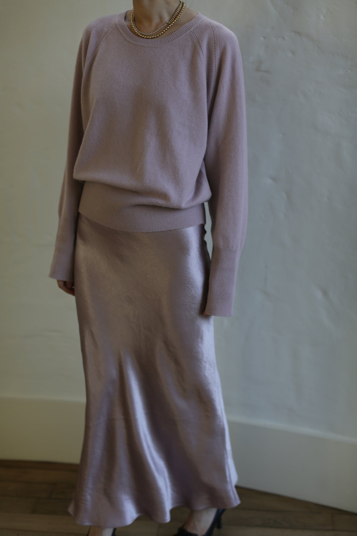 A person stands against a textured white wall wearing a light purple long-sleeve top and matching the Liquid Slip Dress Long | Peony. They are also wearing a gold necklace. The outfit has a soft, elegant appearance.