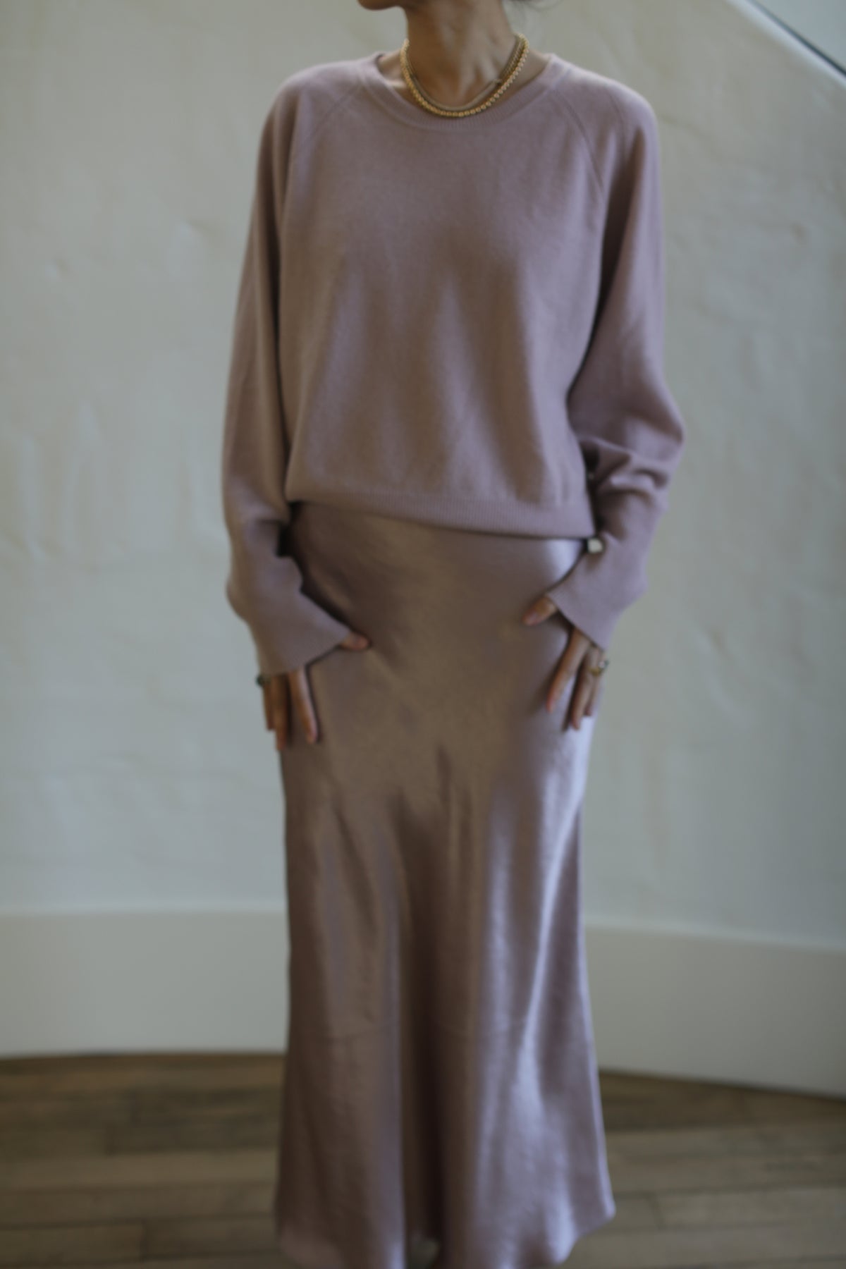 A person is standing indoors wearing a long-sleeved, loose-fitting, Raglan Crewneck Cashmere Sweater | Peony and a matching satin skirt. The background is a plain, light-colored wall, and only the person's torso and part of the legs are visible. They have their hands resting by their sides.