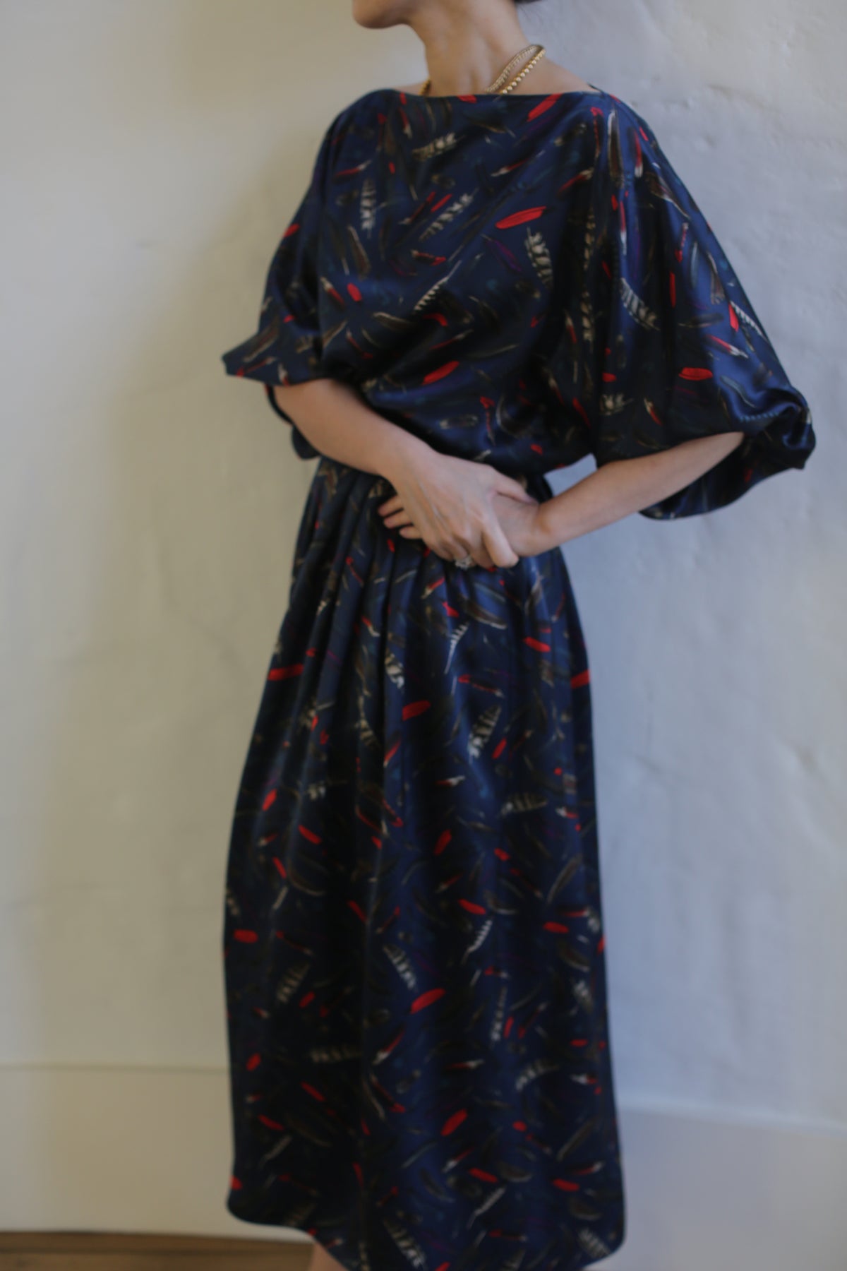 A person stands against a white background wearing a flowing, dark blue Silk Charmeuse Pleated Wrap Skirt | Feather Print Navy with abstract red and beige patterns. The dress features wide, elbow-length sleeves, a gathered waist, and includes a wrap skirt element. The person's hands are clasped in front of them, and their head is not visible in the frame.