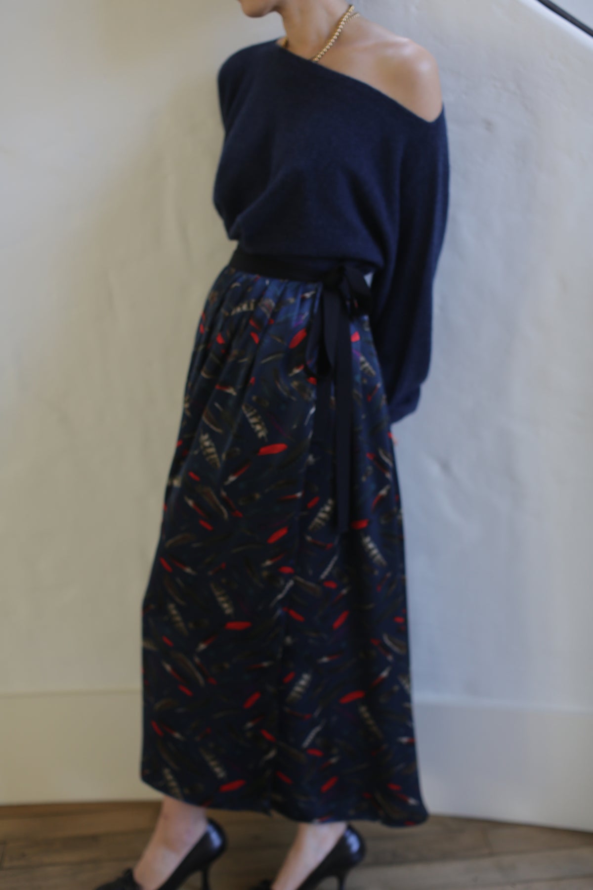 A person is standing against a white background wearing a navy blue sweater and a long, dark Silk Charmeuse Pleated Wrap Skirt | Feather Print Navy. The zero-waste design of the outfit adds an eco-friendly touch. They are also wearing high-heeled shoes. The person’s face is not visible in the image.