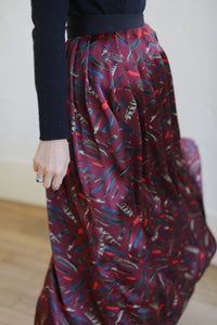 A person in a long-sleeved black top is wearing a Silk Charmeuse Pleated Wrap Skirt | Feather Print Burgundy. The image captures the lower half of their body as they turn gracefully. Their hand is adorned with a ring.