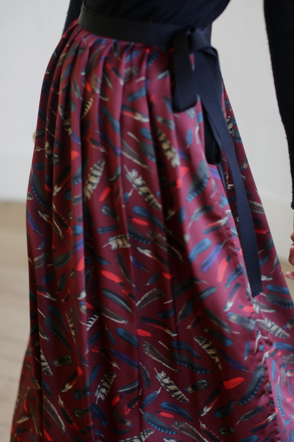 A close-up view of a person wearing a Silk Charmeuse Pleated Wrap Skirt | Feather Print Burgundy. The person is wearing a dark top, and there is a black ribbon tied at the waist of the wrap skirt. The background is out of focus.