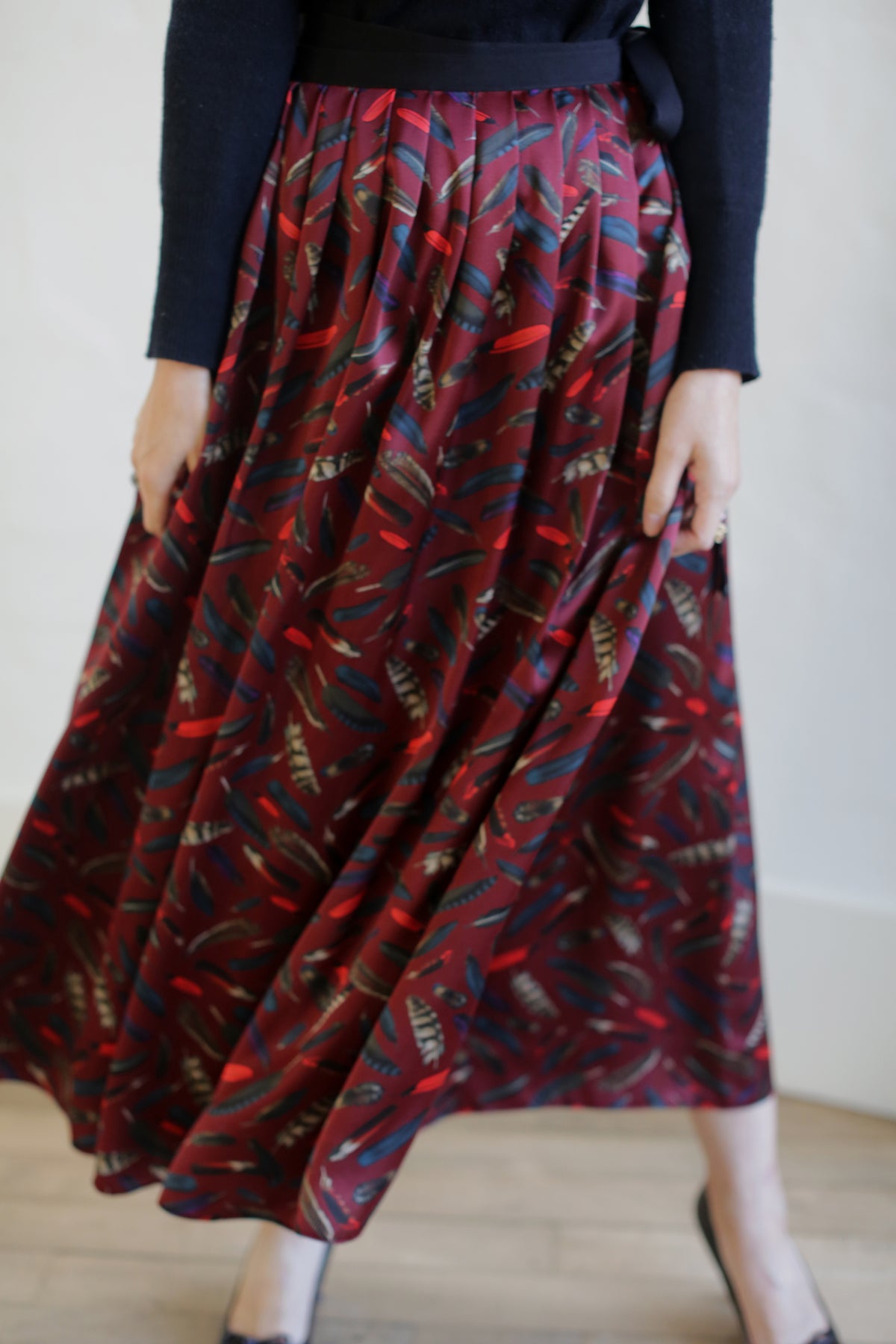 Person wearing a Silk Charmeuse Pleated Wrap Skirt | Feather Print Burgundy. Their upper body is mostly out of the frame, and they are standing against a plain, light-colored background.