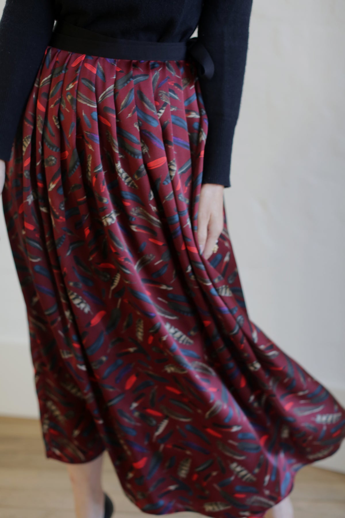 A person is standing in a black long-sleeved top and a Silk Charmeuse Pleated Wrap Skirt | Feather Print Burgundy, which features a flowing, pleated design with abstract motifs in blue, black, and off-white. The image captures the lower half of their body in motion, showcasing the skirt's fluidity.
