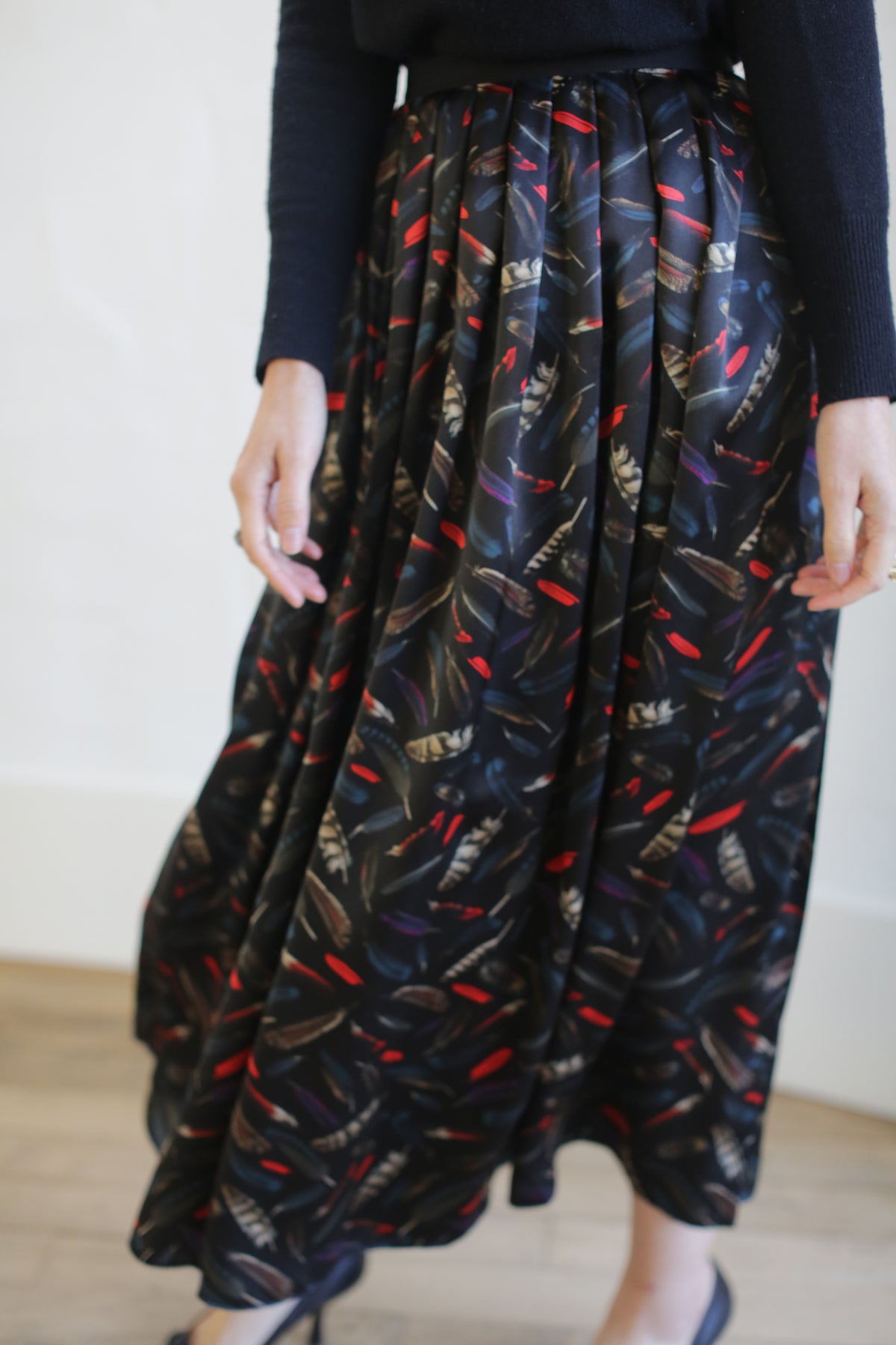 A person wearing a Silk Charmeuse Pleated Wrap Skirt | Feather Print Black with a colorful abstract pattern of feathers and streaks on a black background. The person is also wearing a black top. They are captured from the waist down, standing on a wooden floor.