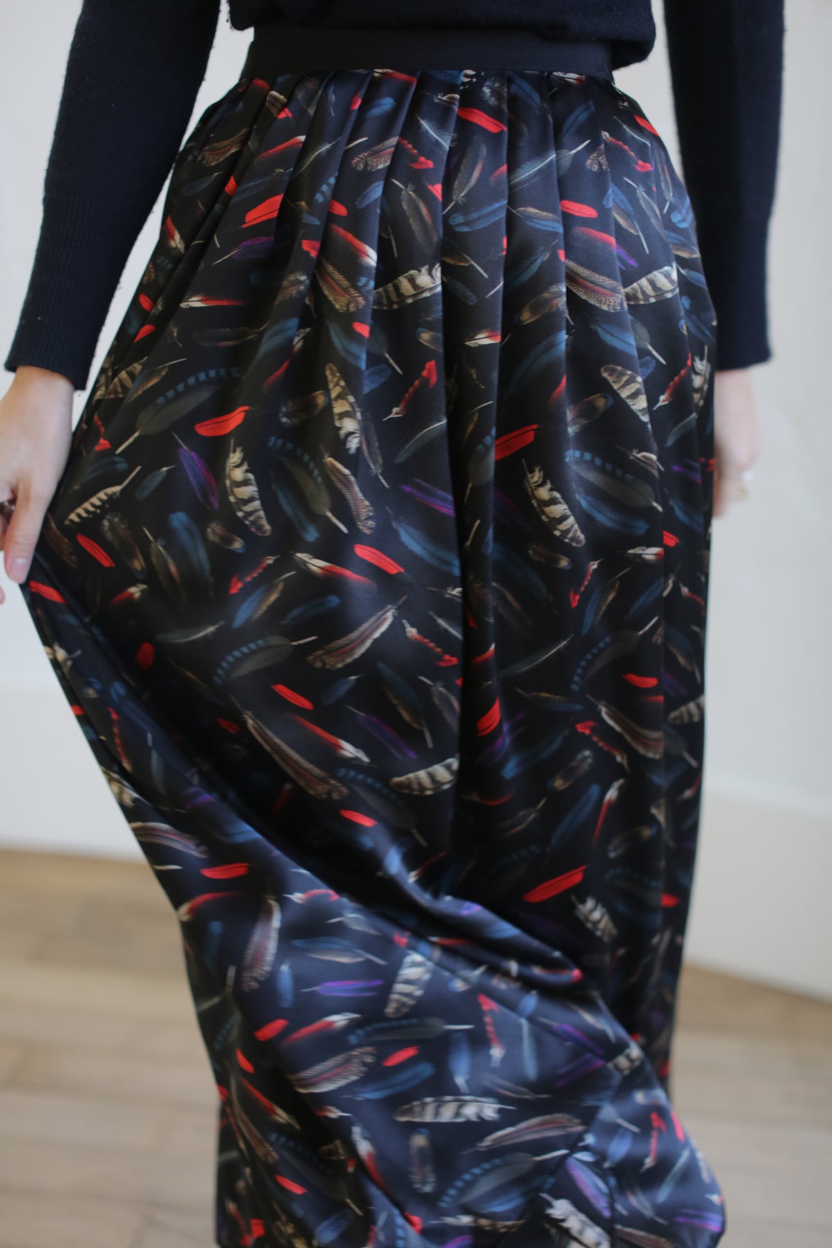 A person is wearing the Silk Charmeuse Pleated Wrap Skirt | Feather Print Black. Crafted from elegant silk charmeuse, the pleated skirt flows gracefully, held slightly out to the side by one hand. They are standing indoors on a wooden floor, with a light-colored wall in the background.