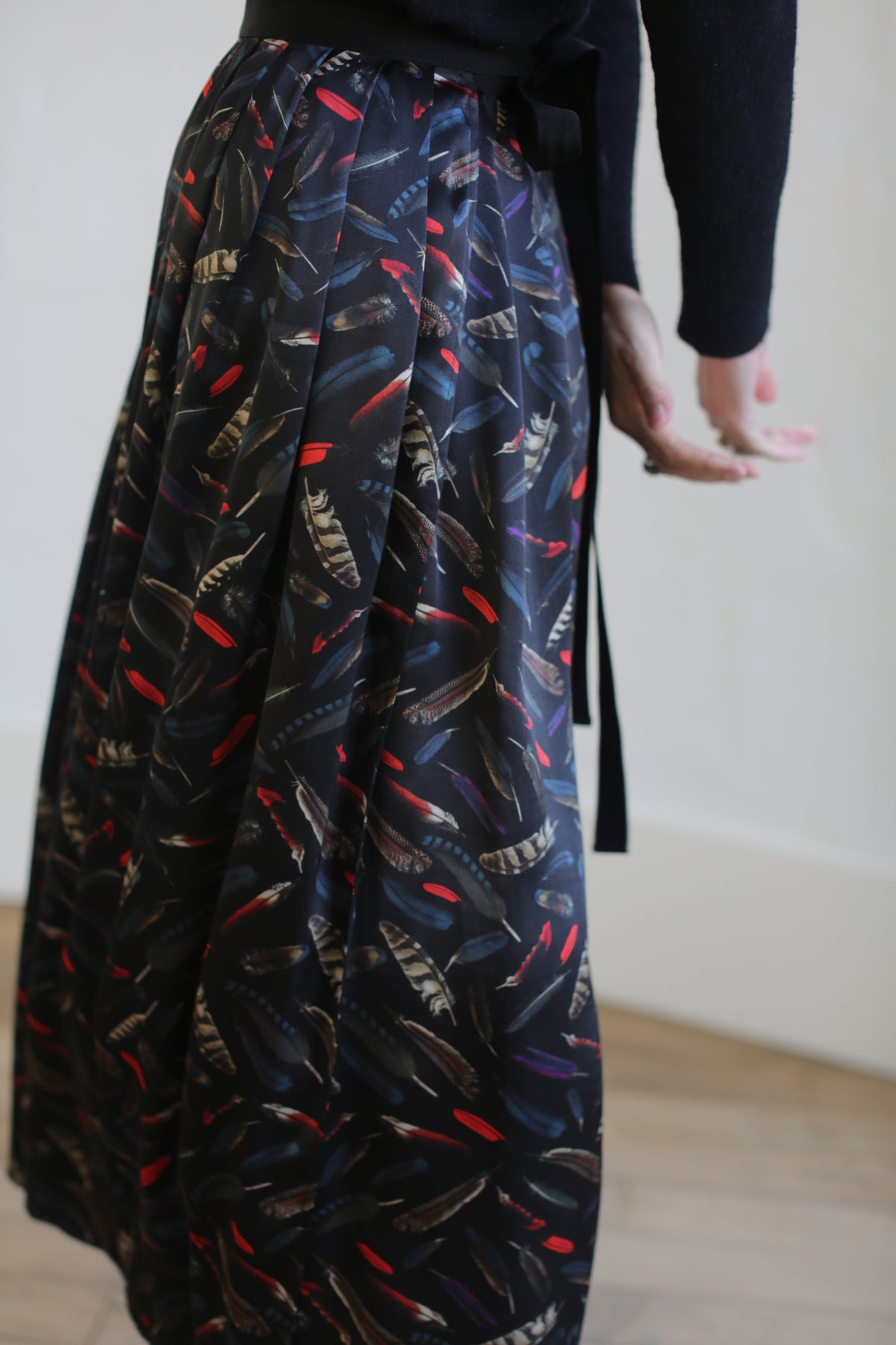 A person wearing a black long wrap skirt with colorful feather patterns stands with one arm slightly extended. The background is a plain white wall and a wooden floor. The focus is on the exquisite Silk Charmeuse Pleated Wrap Skirt | Feather Print Black.