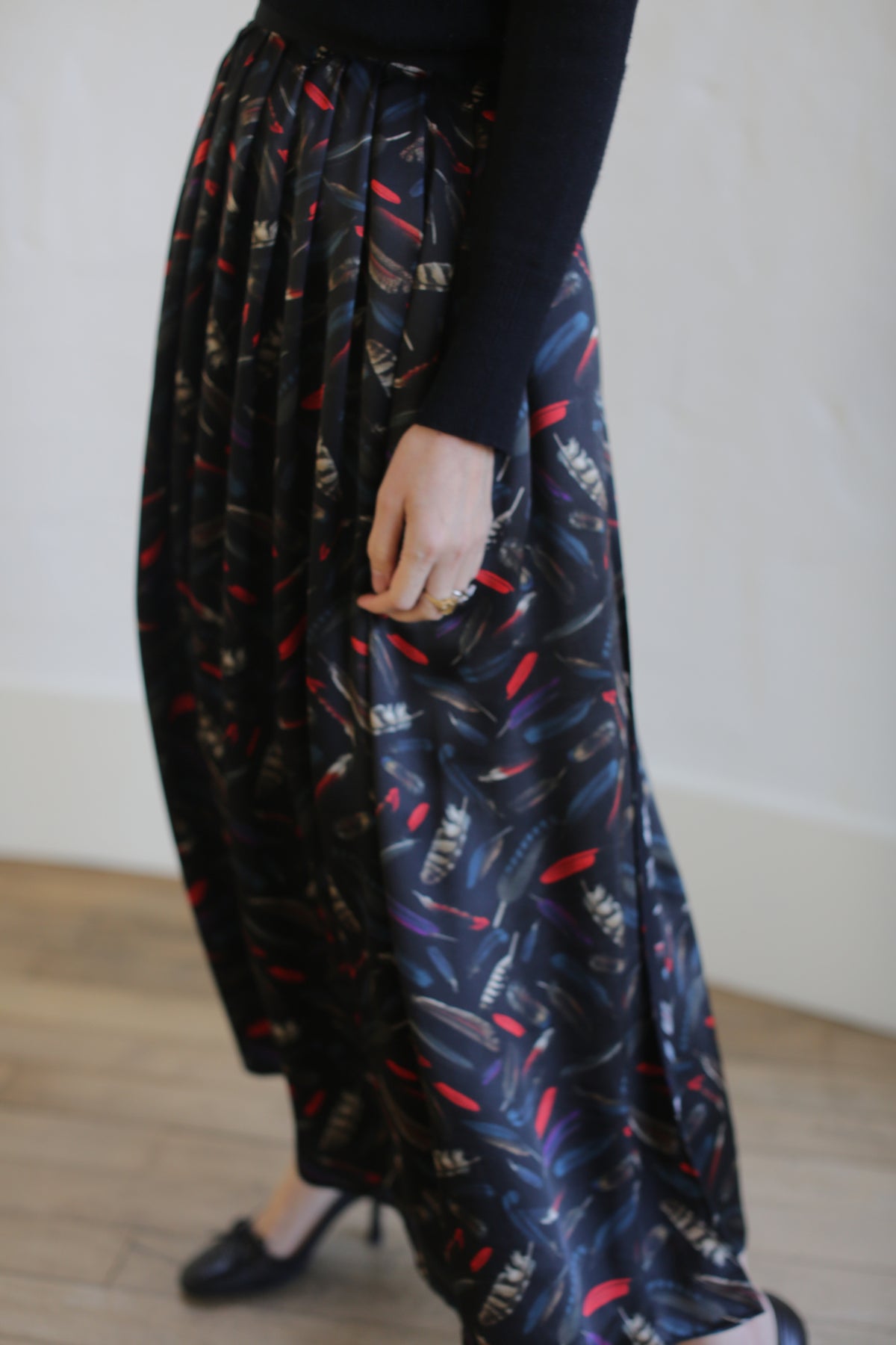 Person wearing a black long-sleeved top and a dark, patterned Silk Charmeuse Pleated Wrap Skirt | Feather Print Black with red and white accents. Only the lower half of the body is visible, including the hand elegantly holding the skirt. The background is a neutral, softly focused indoor setting.