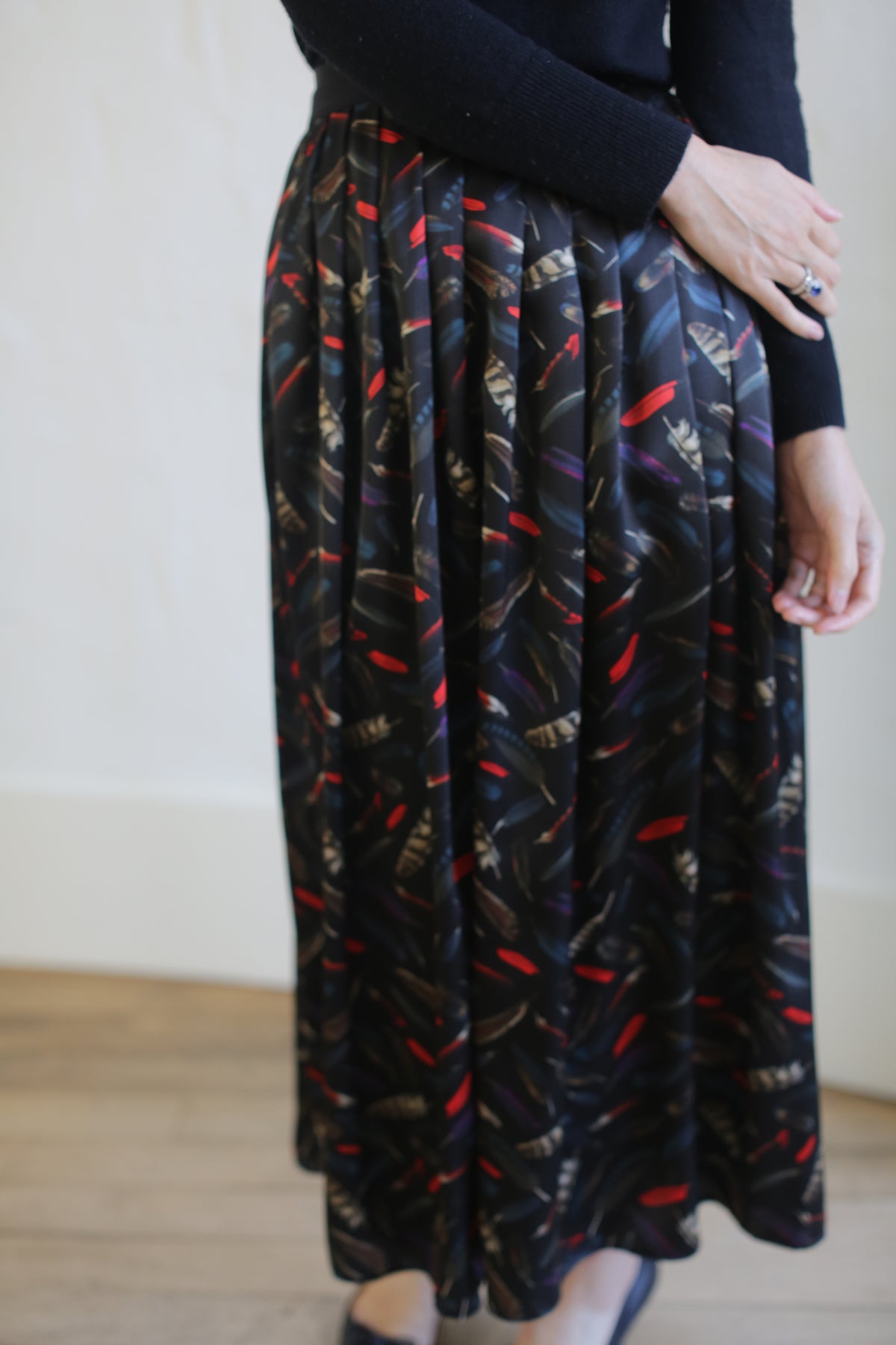 A person wearing a black long-sleeved top and a Silk Charmeuse Pleated Wrap Skirt | Feather Print Black in red, gold, and blue. Their left hand rests on their right arm while standing on a wooden floor against a plain light-colored background.