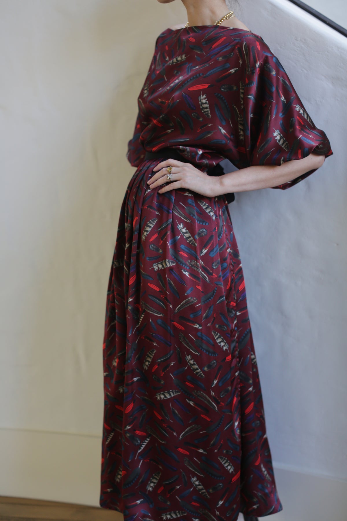 A person is wearing a Silk Charmeuse Pleated Wrap Skirt | Feather Print Burgundy. The skirt features a high waistband and pleated design. The person is standing against a light-colored wall, with one hand resting on their hip.