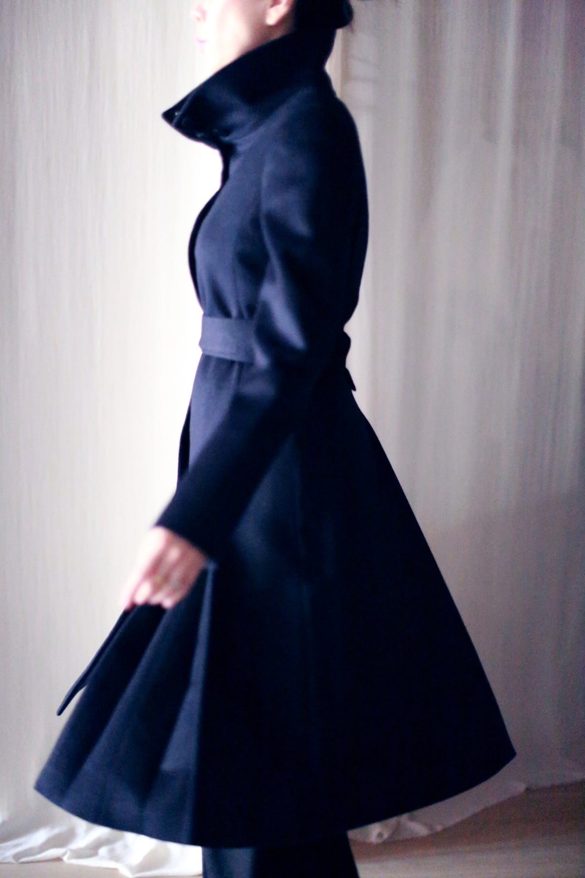 A person wearing the dark and stylish Hyde skirted trench gracefully walks past a backdrop of soft white curtains. The woven cashmere coat with its high collar and belt creates a sleek and elegant silhouette. The person's face is partially obscured, adding an air of mystery to the scene.