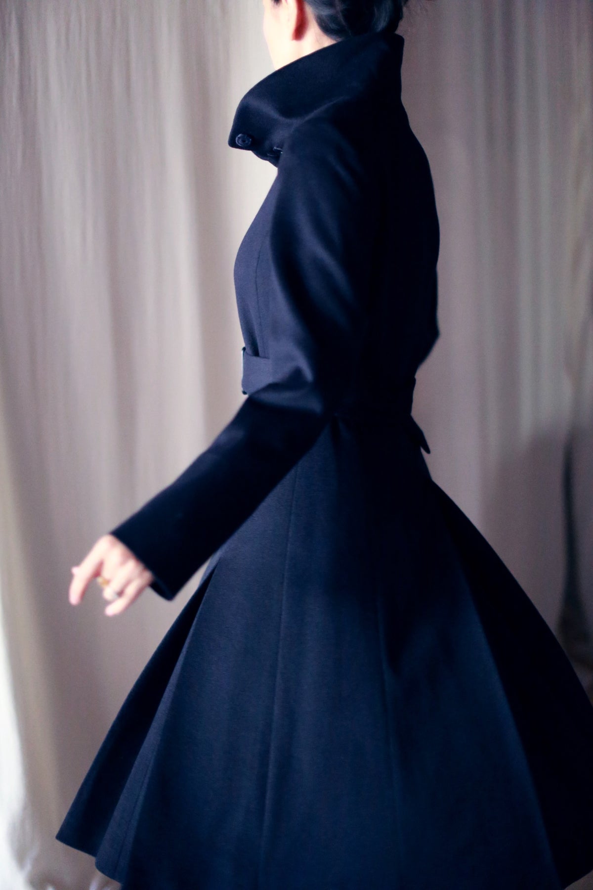A person wearing the Hyde, a long, dark skirted trench with a high collar, poses against a light curtain backdrop. The focus is on the impeccable structure and silhouette of this woven cashmere coat.