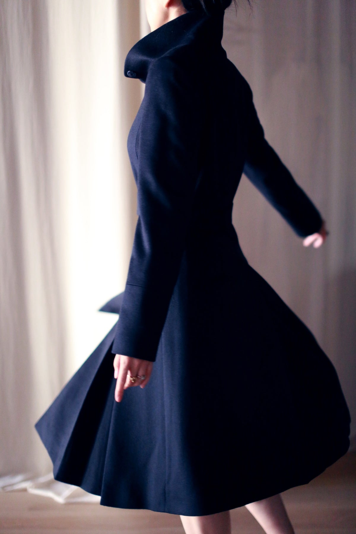 Wearing the Hyde, a person twirls gracefully in this dark, skirted trench coat, creating motion in the fabric. The softly lit background adds an elegant and fluid touch to the scene.