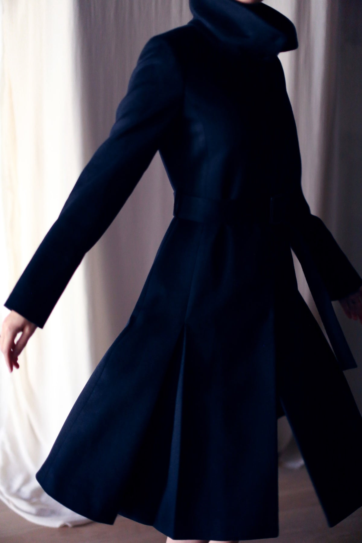 A person wearing the Hyde, a dark and elegant skirted trench with a high collar, spins gracefully. The long sleeves and flared hem are captured mid-movement against a softly lit backdrop.