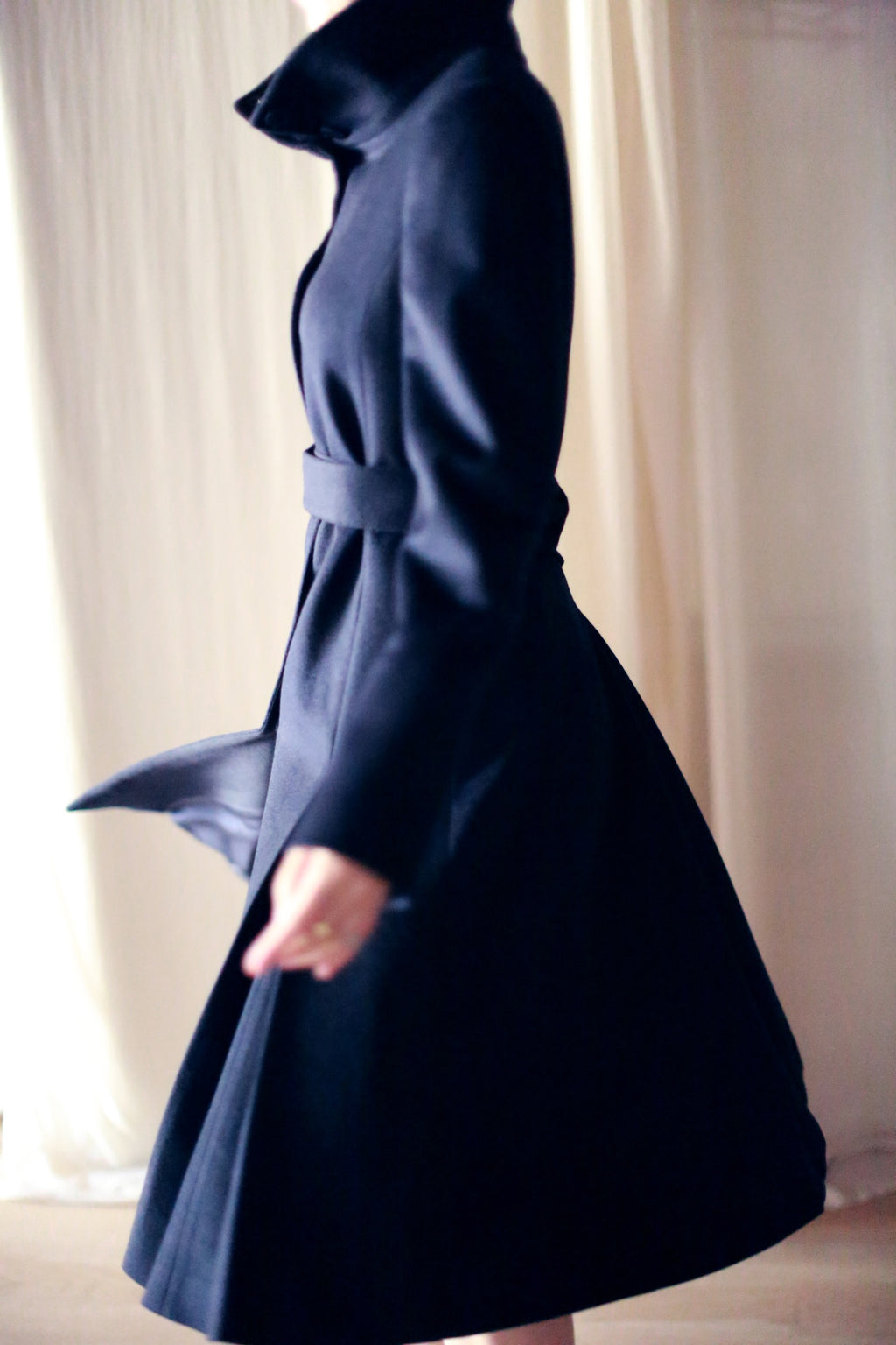 A person wearing the Hyde trench coat, featuring a dark, skirted design with a high collar, is photographed in profile. The fabric of this coat appears to be heavy, woven cashmere. The motion of the person creates a blurred effect against the softly lit background.
