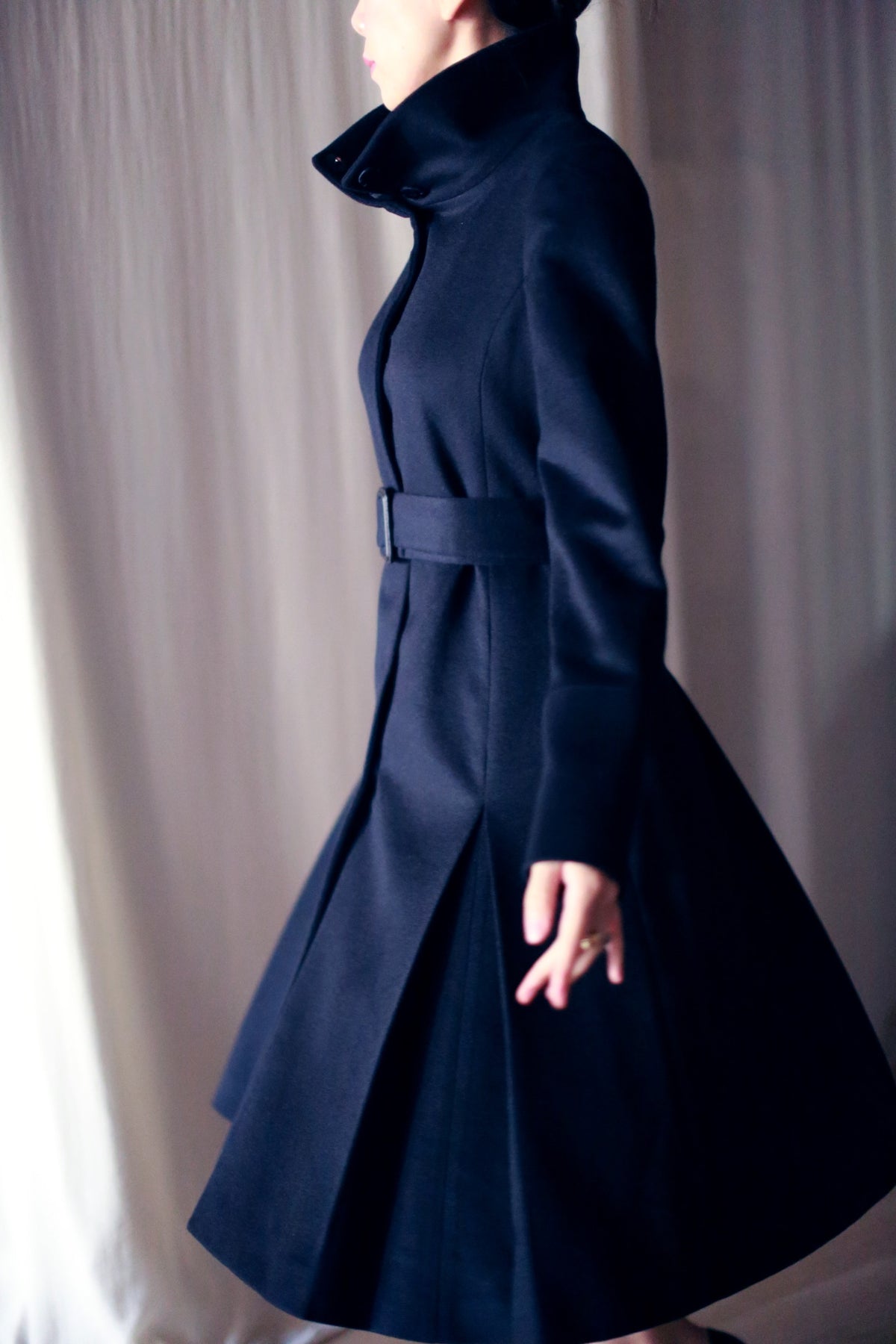 A person in a dark, skirted Hyde trench with a high collar stands against a light background, showcasing their side profile. The coat's woven cashmere flares elegantly, complemented by long sleeves that enhance its refined silhouette.