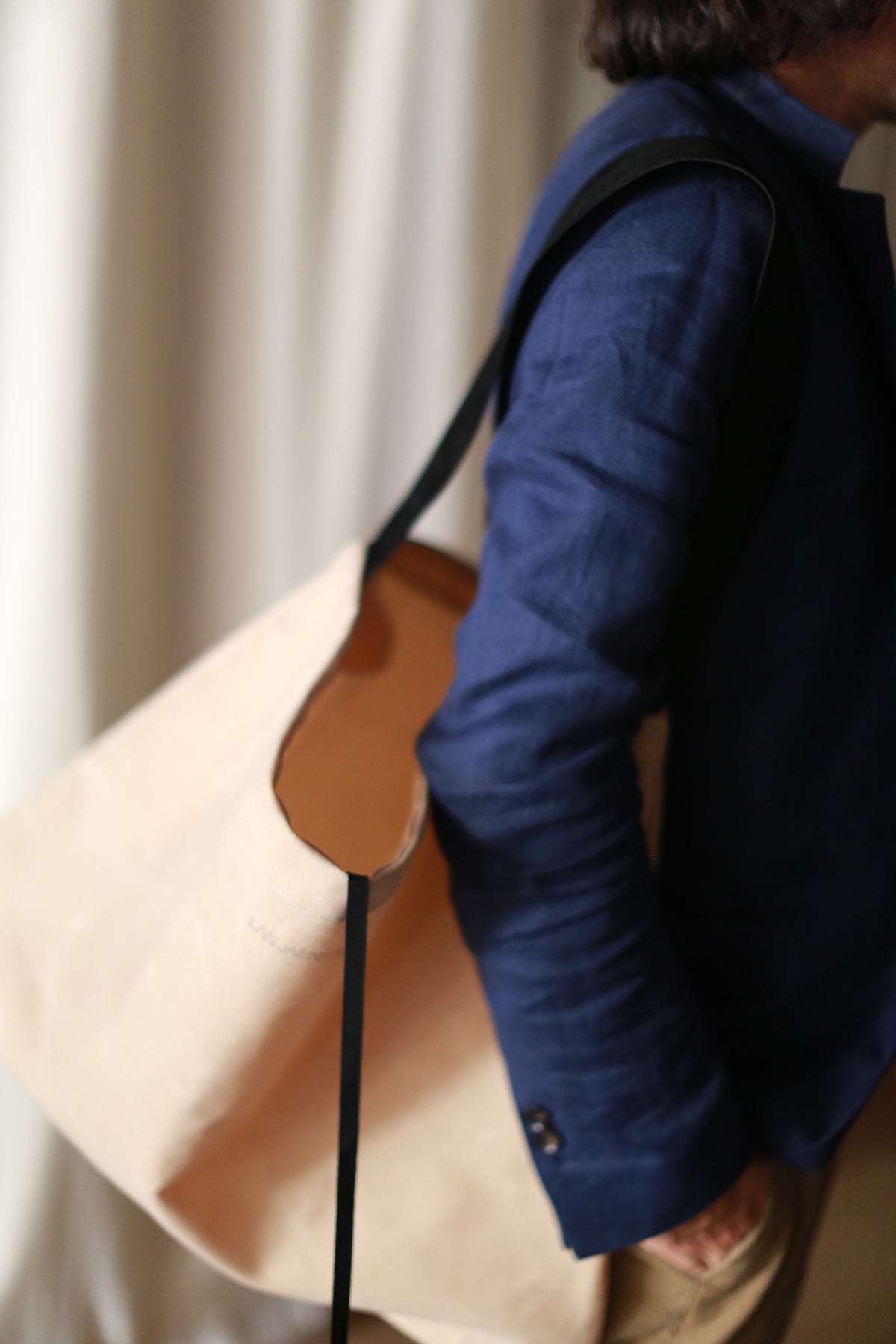 A person wearing a blue long-sleeve shirt and khaki pants carries The Everything Bag, a large hand-made beige tote crafted from cotton canvas with black straps, slung over their shoulder. The background is blurred.