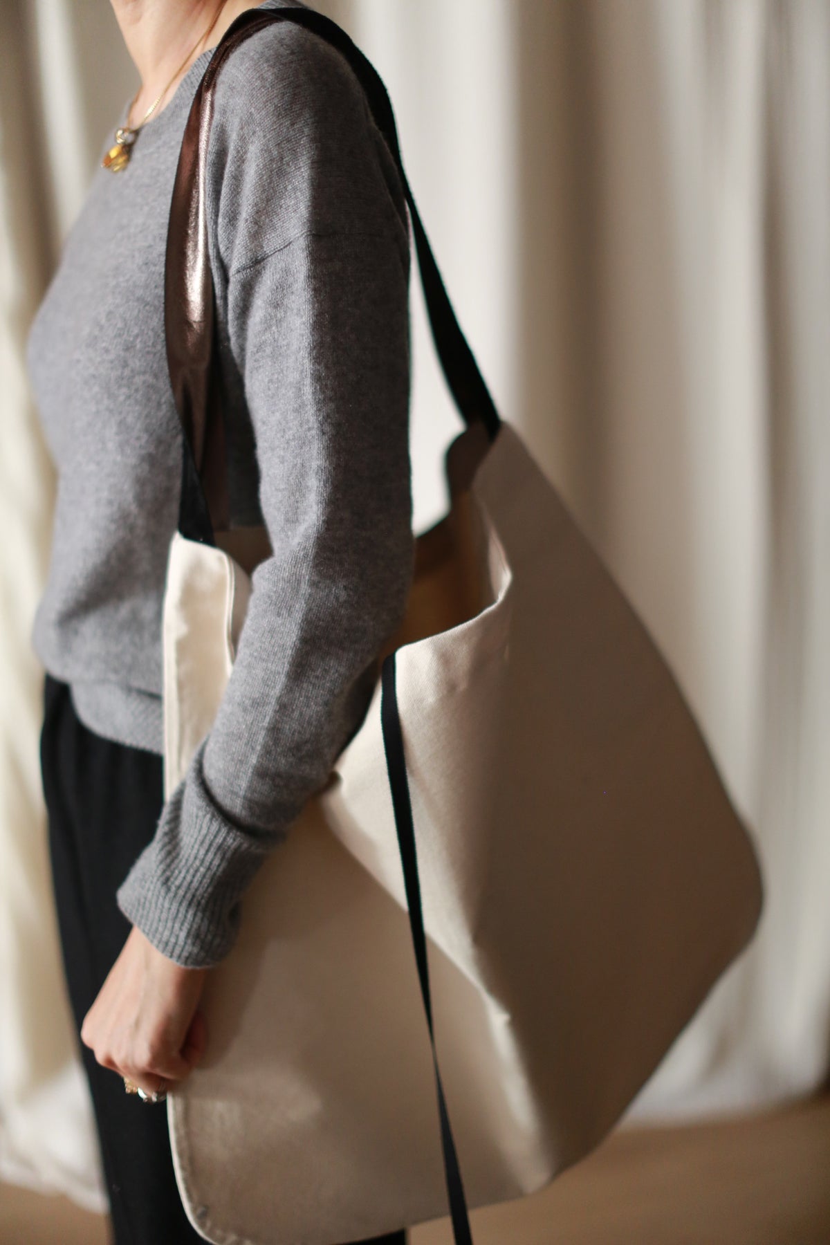 A person wearing a gray sweater and dark pants is carrying The Everything Bag, a large, hand-made tote crafted from soft Napa lambskin with black straps, over their shoulder. The background is blurred, drawing attention to the person and the exquisite bag.