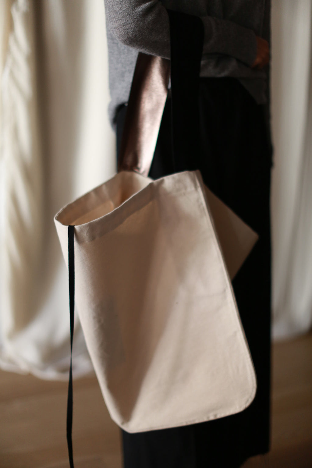 A person stands indoors, partially in view, carrying The Everything Bag—a large, hand-made beige canvas tote bag—over their shoulder. They are dressed in a grey top and black pants, with soft, blurred draped fabric creating a calm and neutral-toned background.