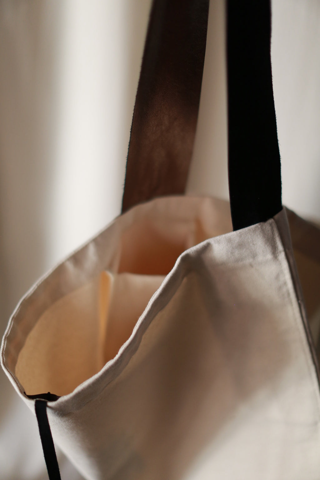 A close-up of The Everything Bag, a hand-made tote crafted from beige cotton canvas, featuring dark brown and black handles. The bag appears empty, and the light is soft, creating a gentle, unfocused background. The open top of the bag and its simple design are highlighted.