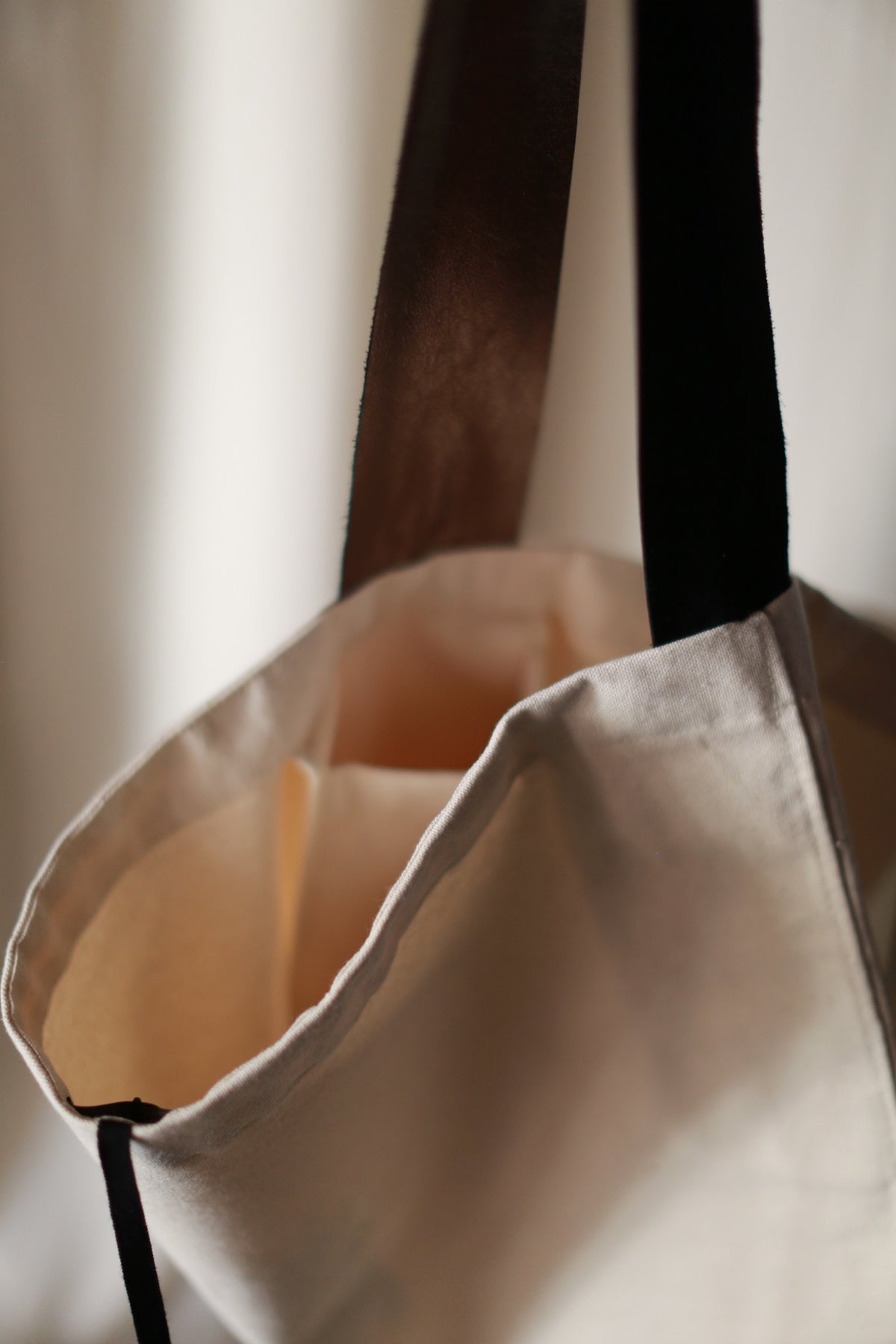 A close-up of The Everything Bag, a hand-made tote crafted from beige cotton canvas, featuring dark brown and black handles. The bag appears empty, and the light is soft, creating a gentle, unfocused background. The open top of the bag and its simple design are highlighted.
