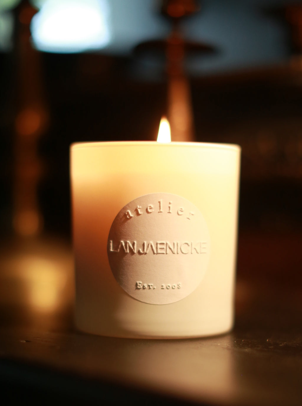 A lit House Candle, with an elegant white design, sits on a dark surface. The candle features an embossed label that reads "Atelier Lan Jaenicke Est. 2008" and emits a delicate fragrance of subtle woody undertones. The background is softly blurred, showcasing warm tones and indistinct shapes, enhancing the cozy ambiance.