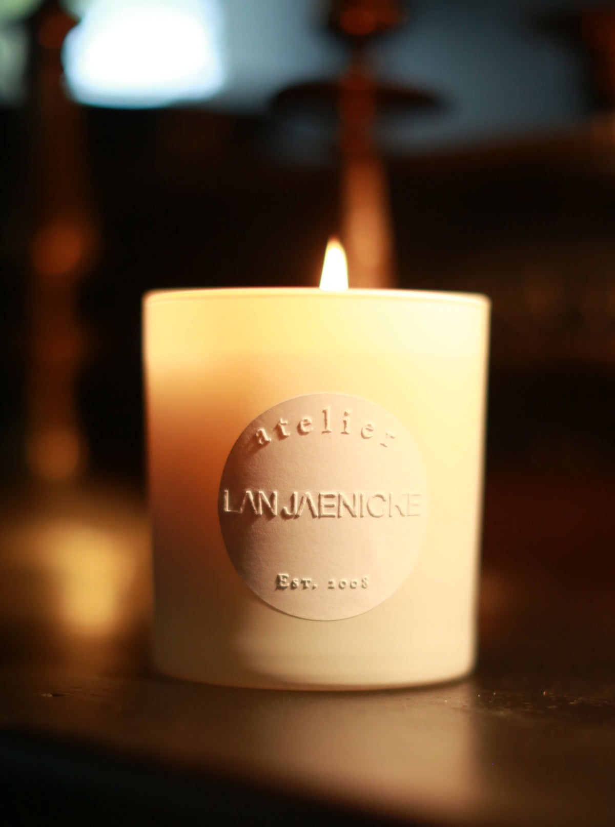 A lit House Candle, with an elegant white design, sits on a dark surface. The candle features an embossed label that reads "Atelier Lan Jaenicke Est. 2008" and emits a delicate fragrance of subtle woody undertones. The background is softly blurred, showcasing warm tones and indistinct shapes, enhancing the cozy ambiance.