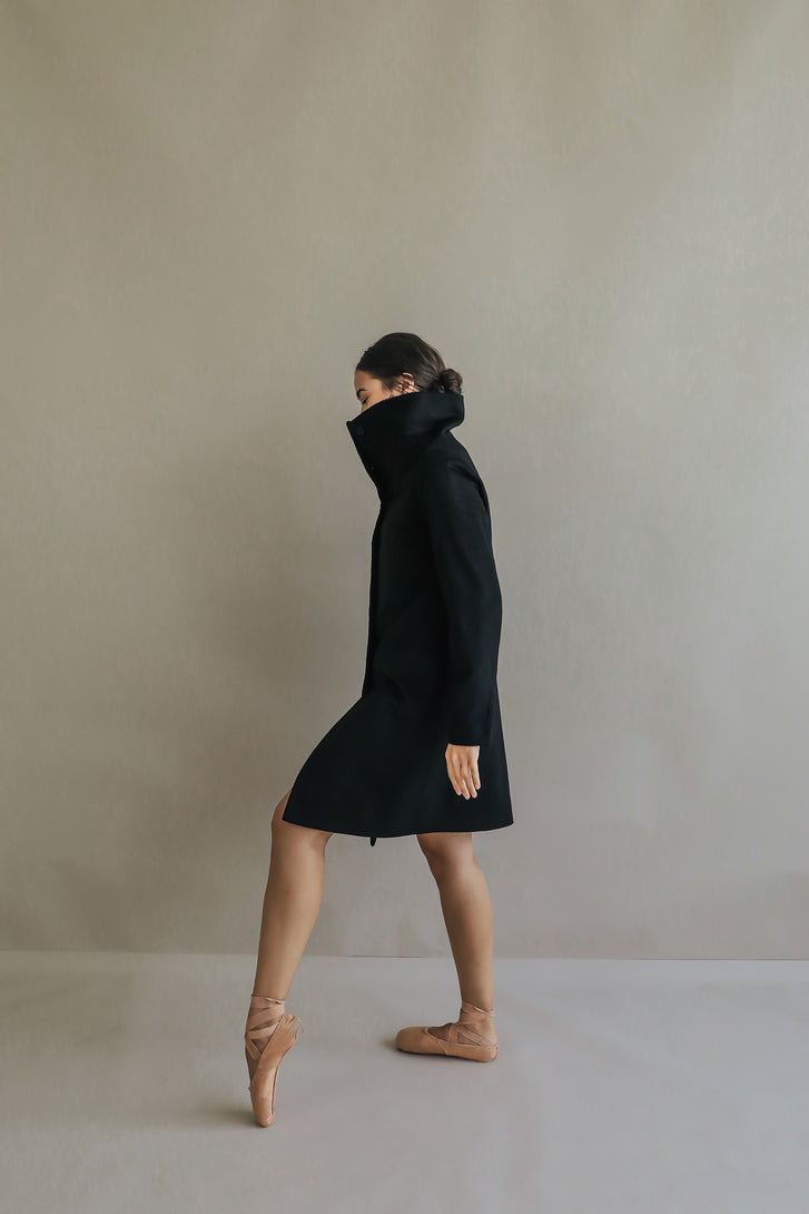 Dressed in the Stamford Long, a person in ballet shoes stands gracefully on tiptoe against a neutral background. The high collar of the cashmere coat partially obscures their face, adding an air of mystery to the elegant pose.