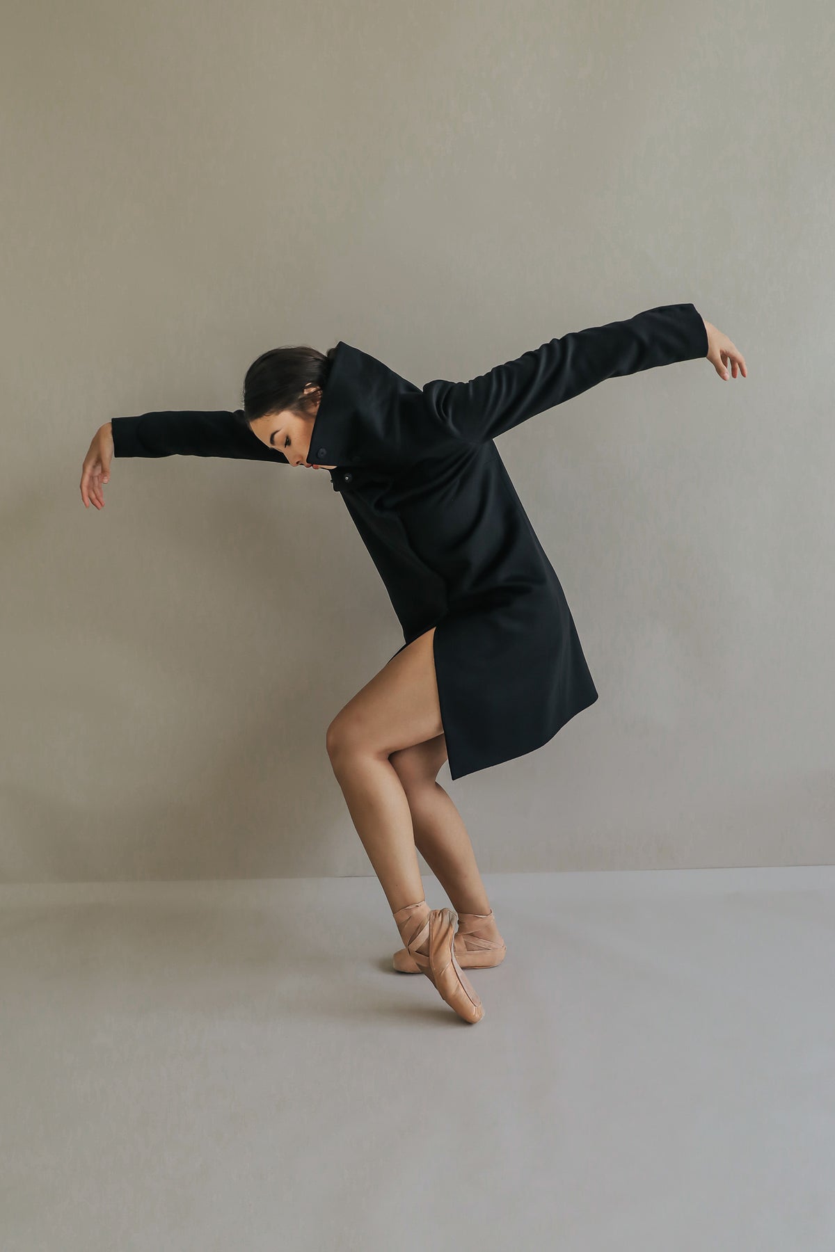 Wearing the Stamford Long, a dancer gracefully poses against a plain background. She balances on one foot with arms extended, exuding elegance and fluidity. Her focused expression and the coat's soft fabric highlight her skill and artistic expression.