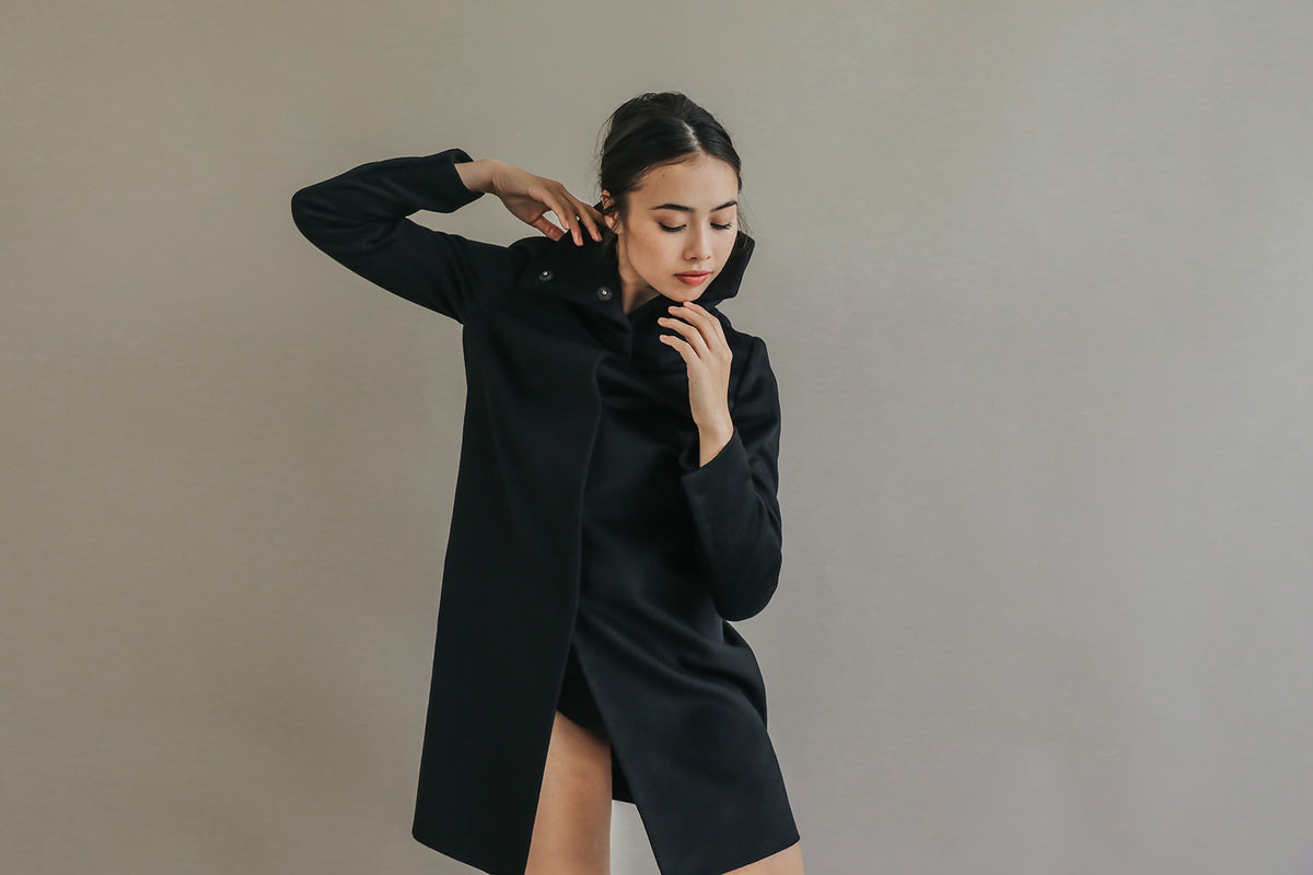 A person poses gracefully against a neutral background, wearing the elegant Stamford Long black cashmere coat with a high collar. One hand is near their face and the other is raised, as they gaze downward thoughtfully.