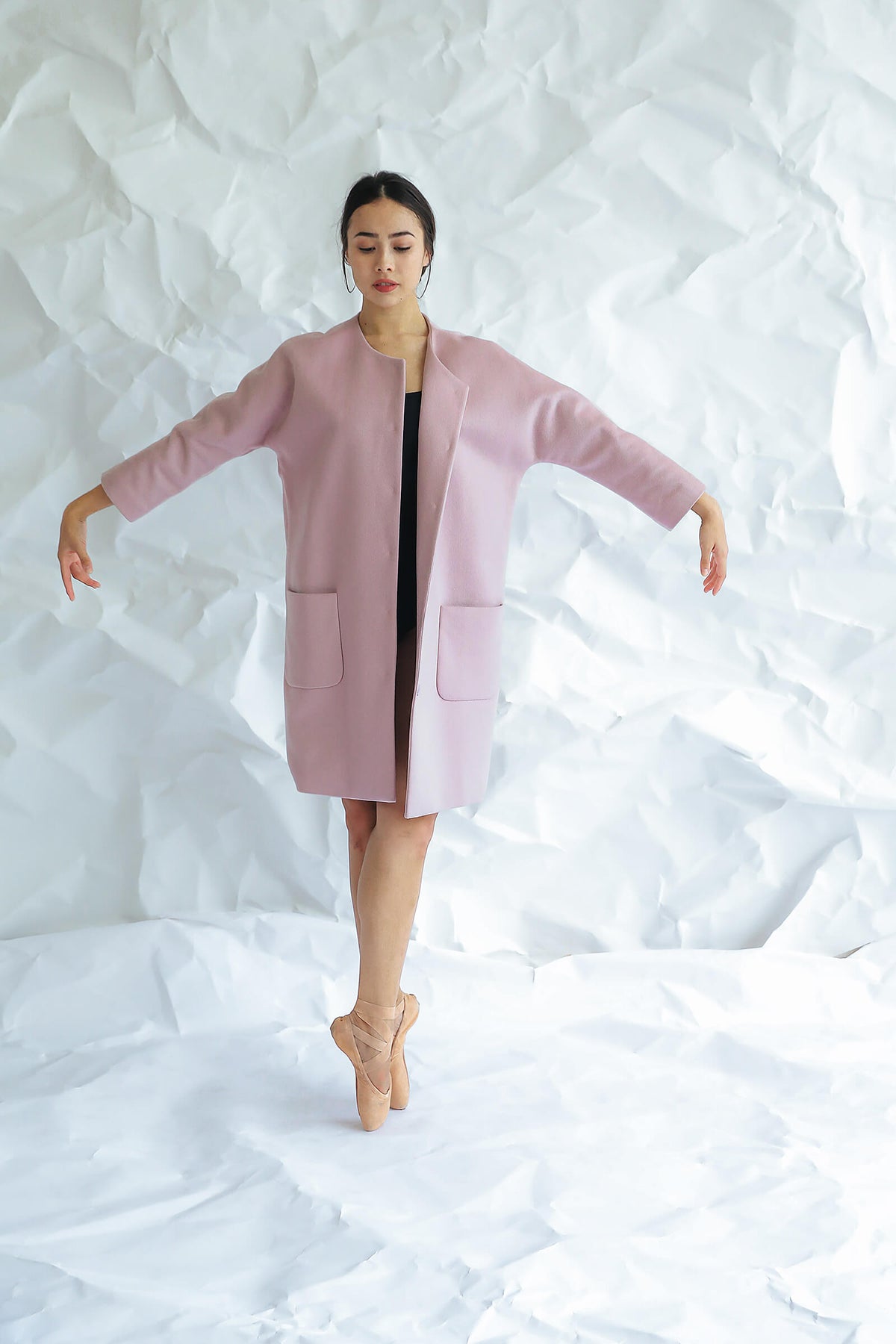 A woman in the Battersea light pink cashmere coat with raglan sleeves and ballet shoes stands en pointe, arms gracefully extended like a ballerina against a crumpled white paper background.