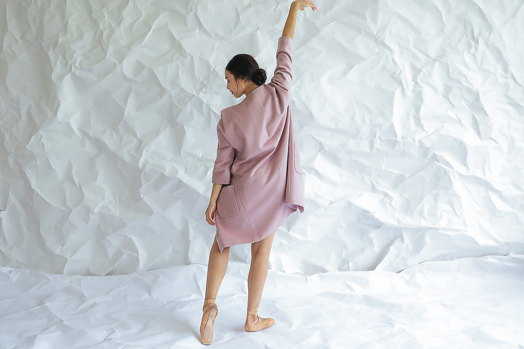 Wearing the Battersea, a loose mauve dress with handmade snap buttons, a woman in ballet shoes balances on one foot with her arm raised, posing gracefully with her back to the camera against a white crumpled paper backdrop.