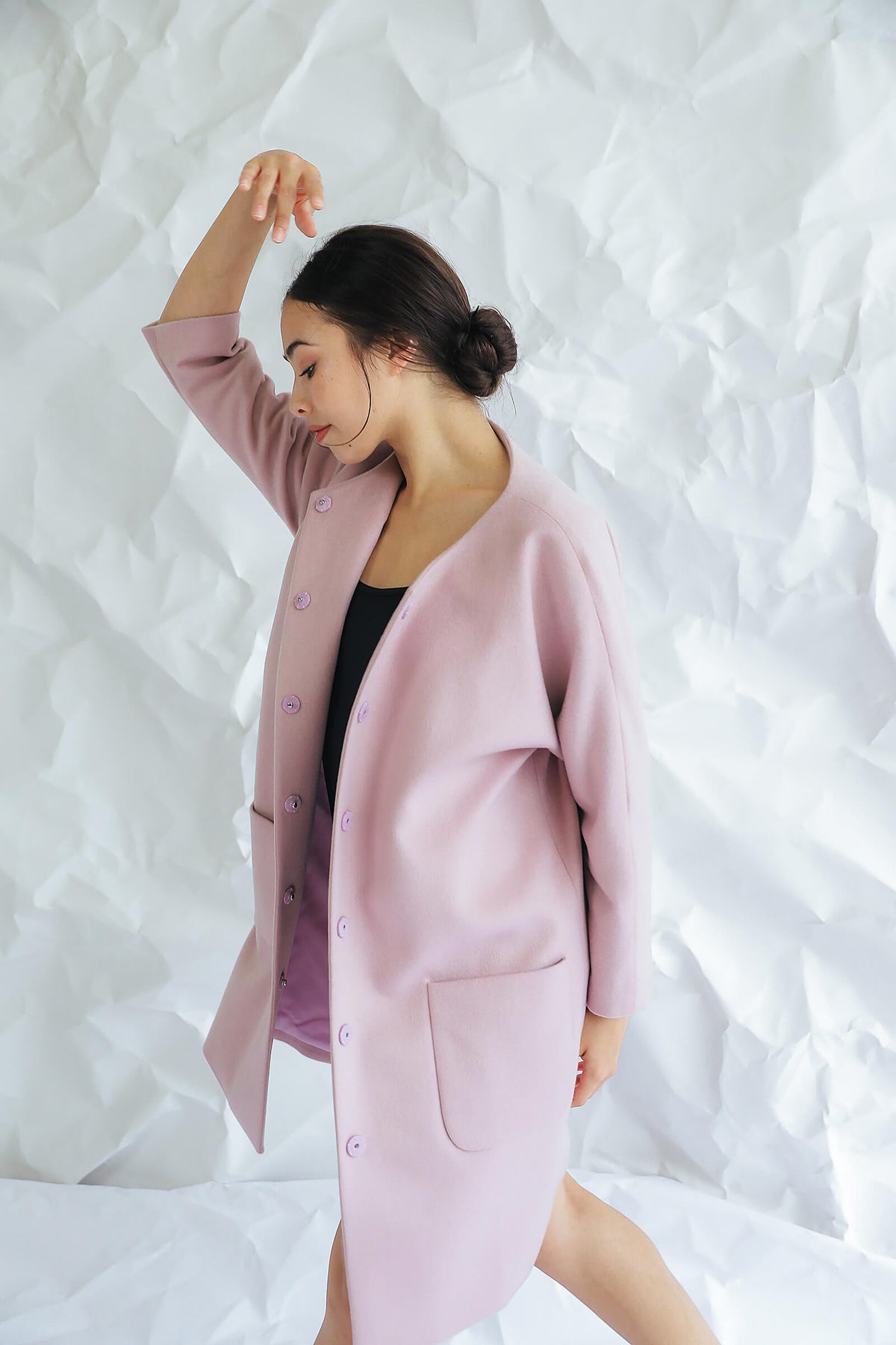 A woman models the Battersea light pink cashmere coat with raglan sleeves, gracefully raising one arm against a crumpled white paper backdrop.