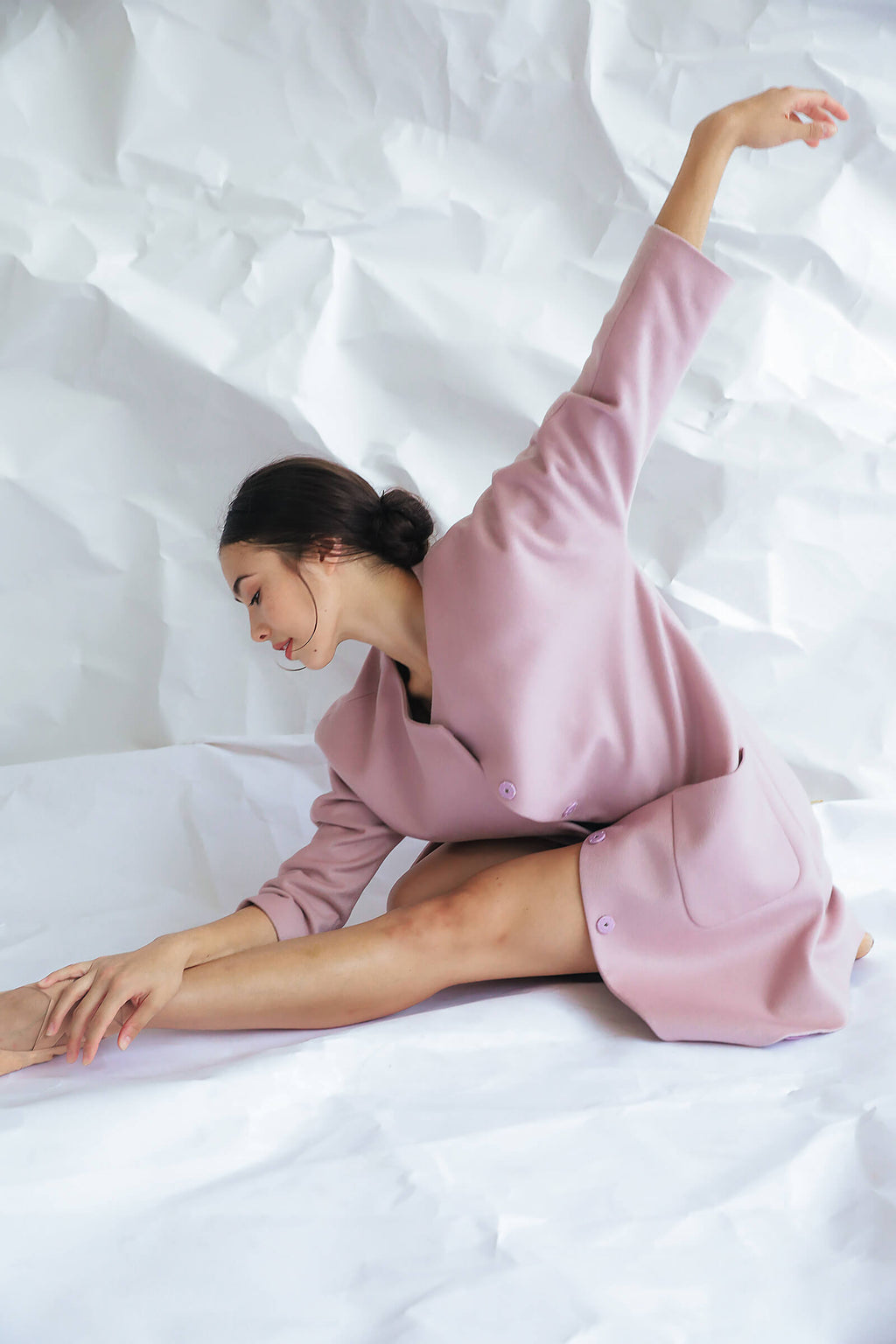 A woman in the Battersea pale pink cashmere coat with raglan sleeves sits on the floor, one leg extended and reaching toward her foot, while her other arm stretches gracefully overhead against a crumpled white backdrop.