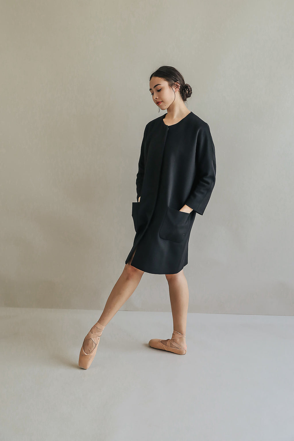 A woman wearing the Battersea black cashmere coat with raglan sleeves and ballet slippers stands on pointe, hands in pockets, looking down against a neutral background.