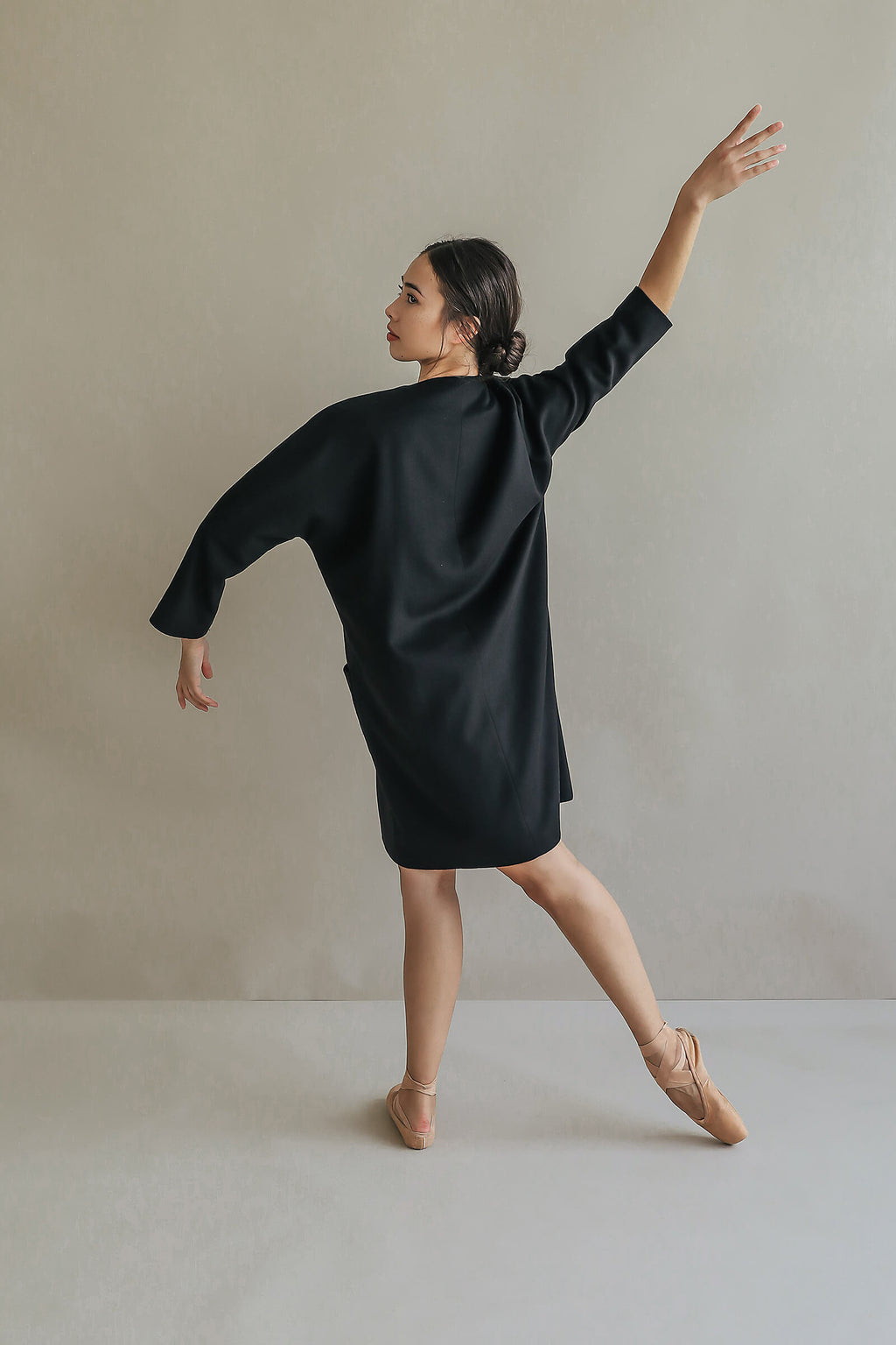 A woman models the Battersea, a loose black dress with raglan sleeves, paired with ballet slippers. She poses gracefully en pointe against a neutral background, one arm raised and the other extended to the side.