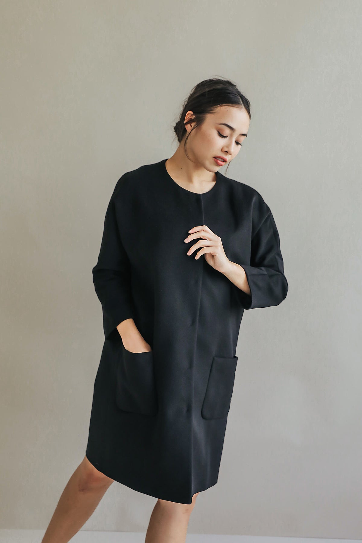 A woman with dark hair tied back wears the Battersea—a loose black dress with raglan sleeves and large front pockets—standing against a plain light gray background, looking down and softly touching her hand to her chest.