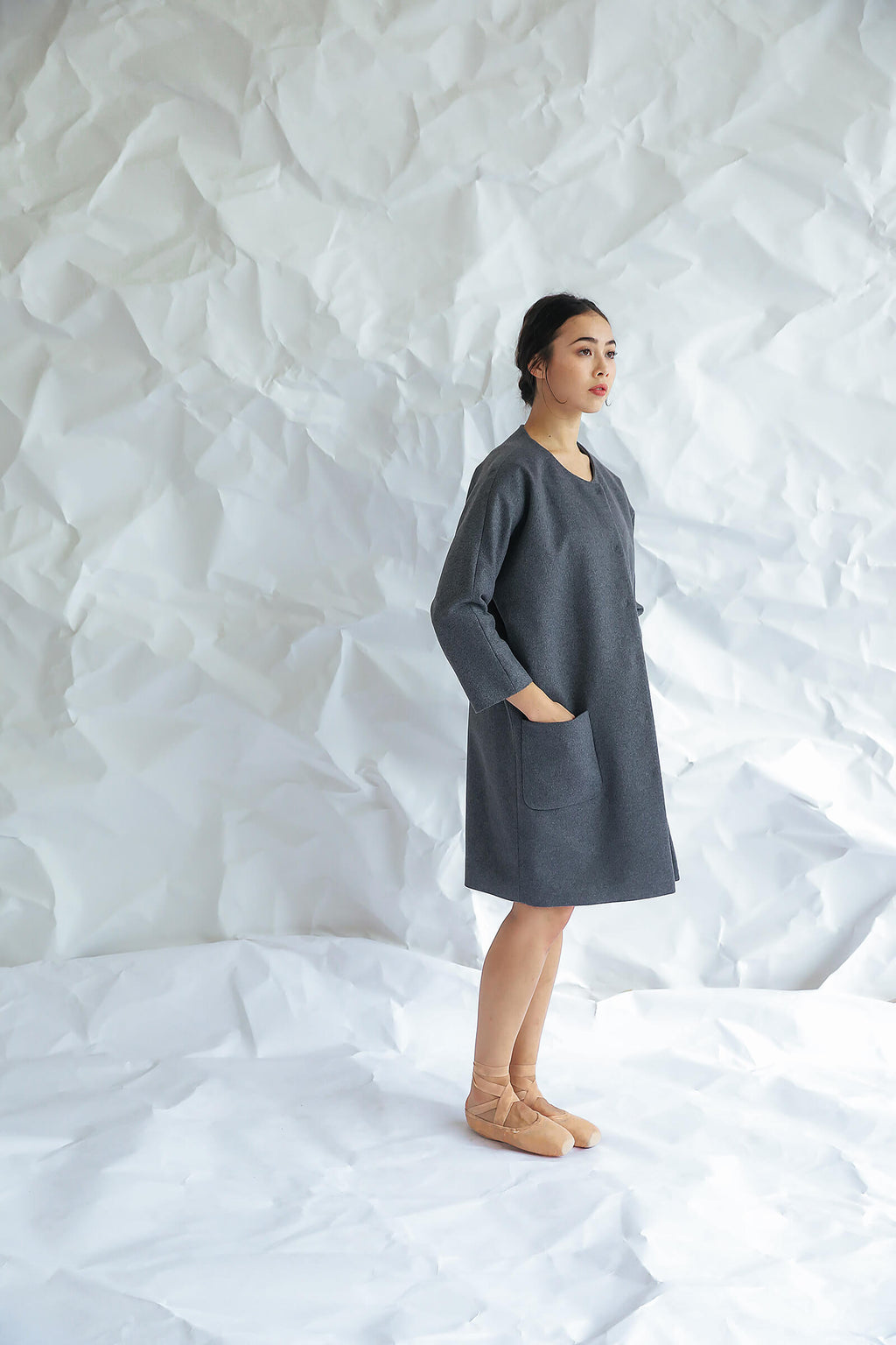 A woman stands sideways on a crumpled white paper background, wearing the Battersea—a loose-fitting gray dress with raglan sleeves and pockets—paired with tan ballet flats, looking forward with a neutral expression.