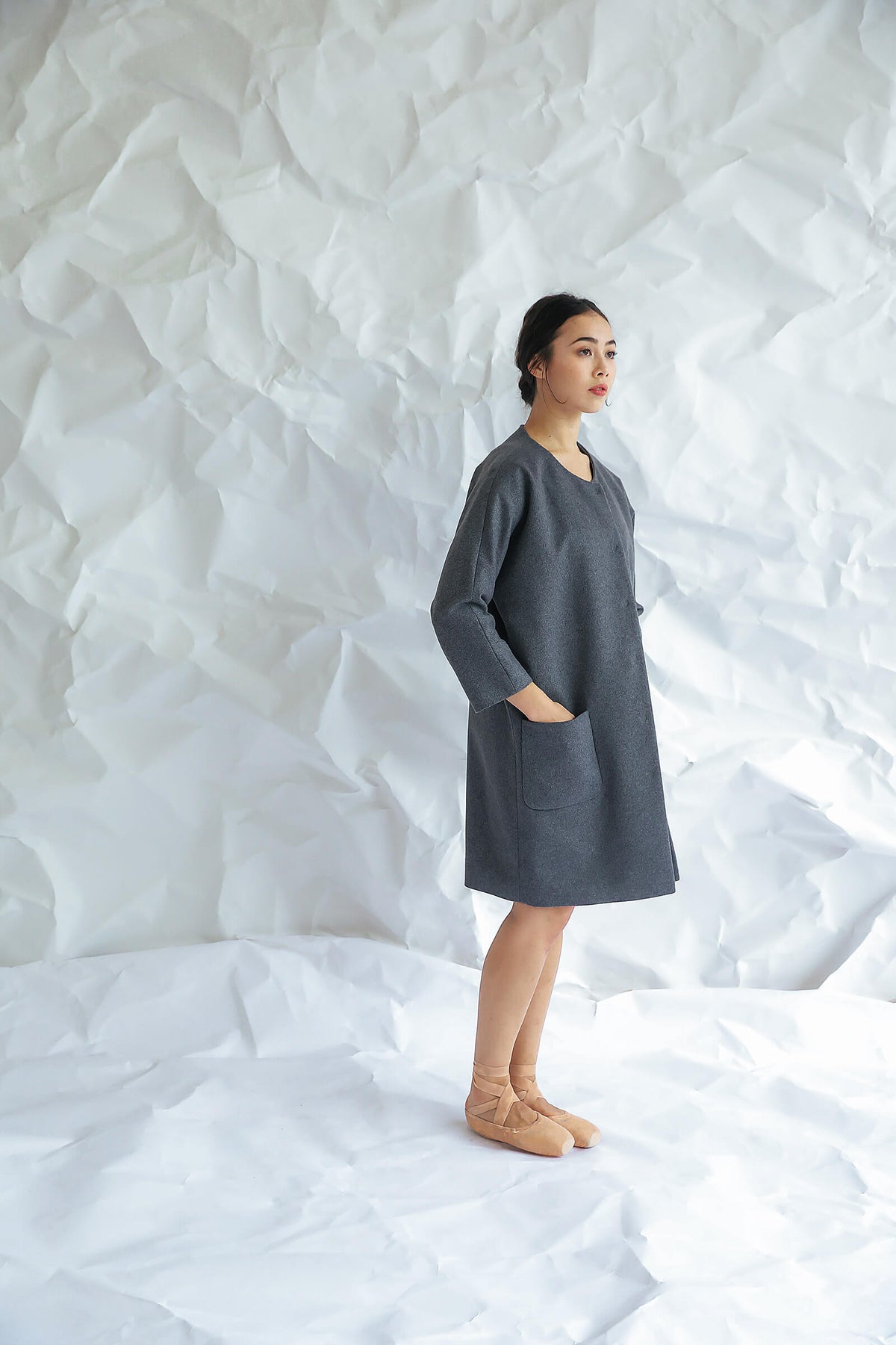 A woman stands sideways on a crumpled white paper background, wearing the Battersea—a loose-fitting gray dress with raglan sleeves and pockets—paired with tan ballet flats, looking forward with a neutral expression.