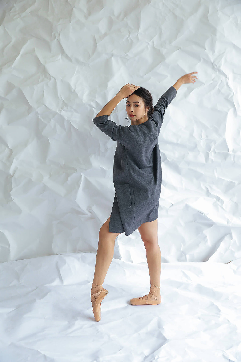 A woman poses gracefully on tiptoe in the Battersea dress— a loose gray style with raglan sleeves—paired with ballet shoes, set against a crumpled white paper backdrop.