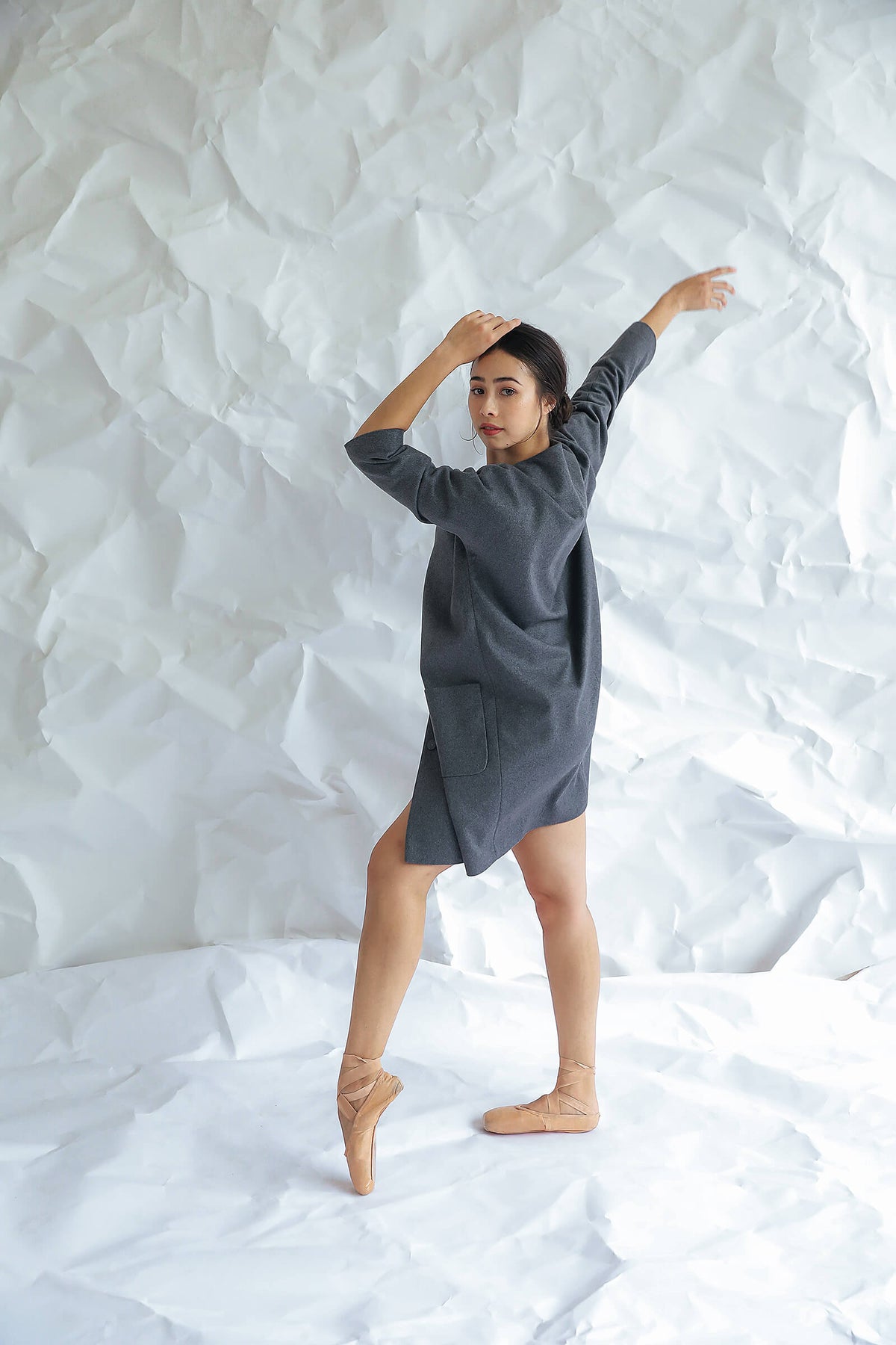 A woman poses gracefully on tiptoe in the Battersea dress— a loose gray style with raglan sleeves—paired with ballet shoes, set against a crumpled white paper backdrop.